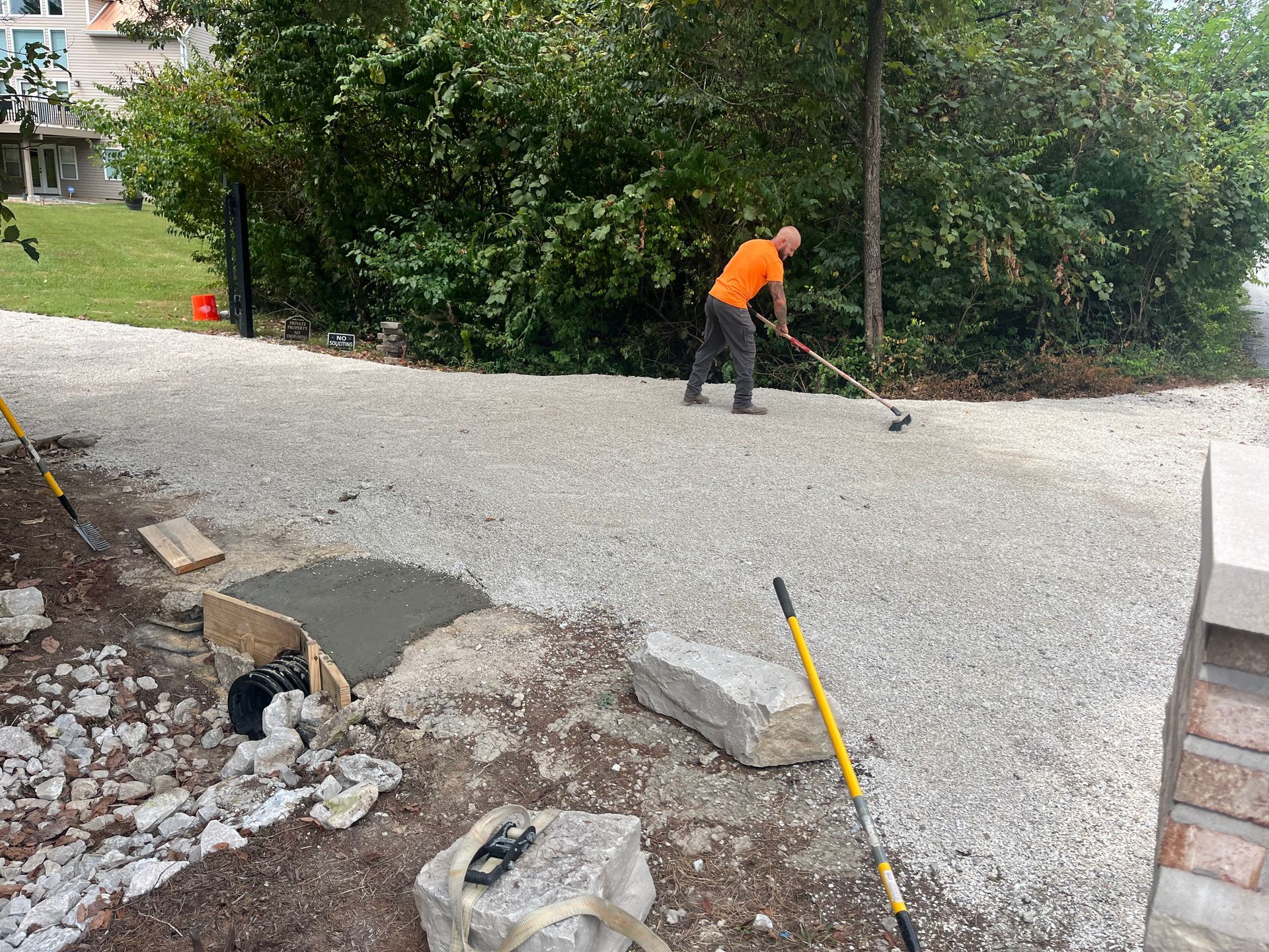 Worker smoothing a gravel driveway beside bushes and construction tools