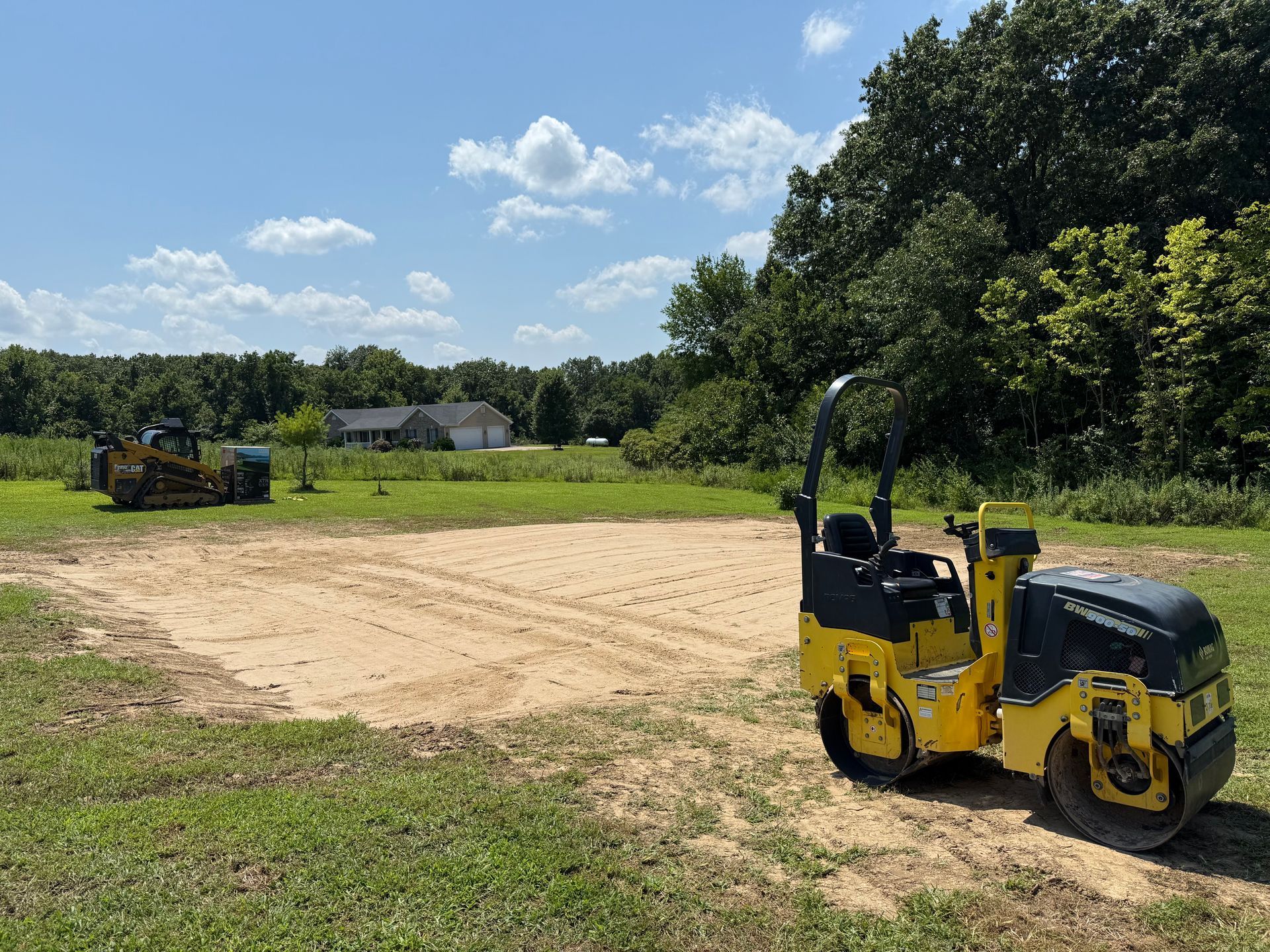 Yellow road roller on a dirt lot near trees under a blue sky