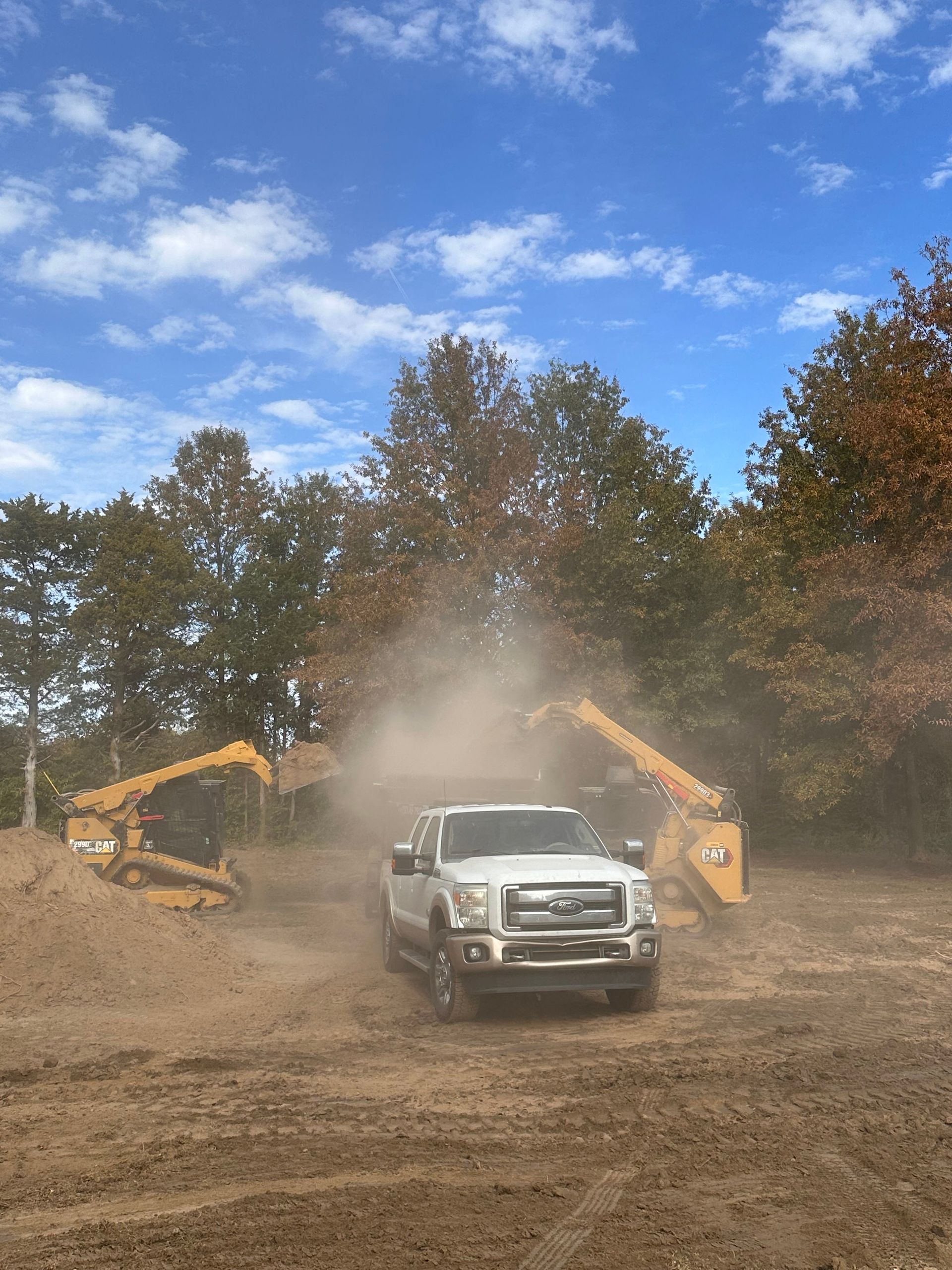 White pickup truck drives through a dusty dirt lot between two yellow excavators under a blue sky.