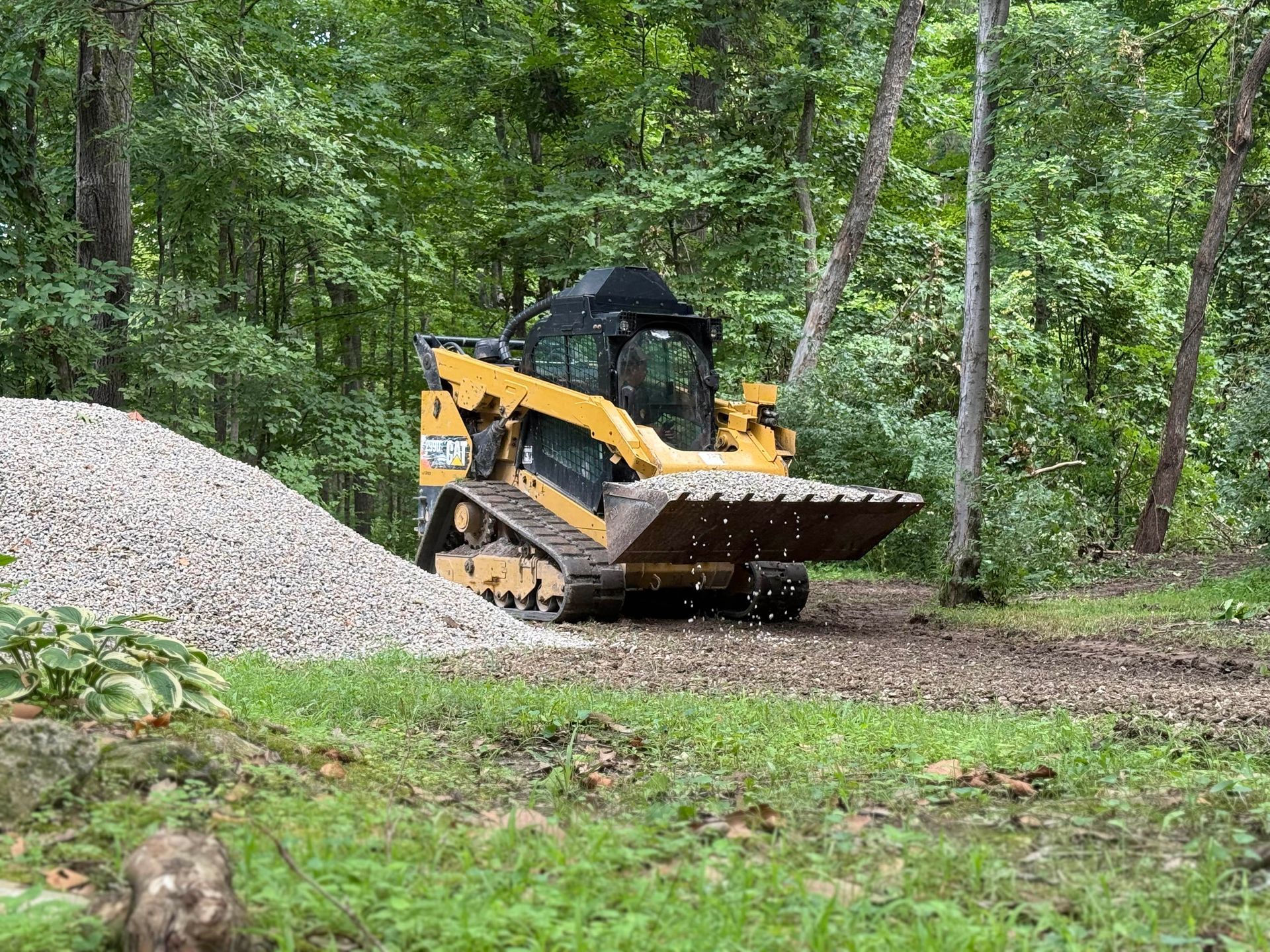 Yellow skid-steer loader driving on a muddy forest path beside a gravel pile