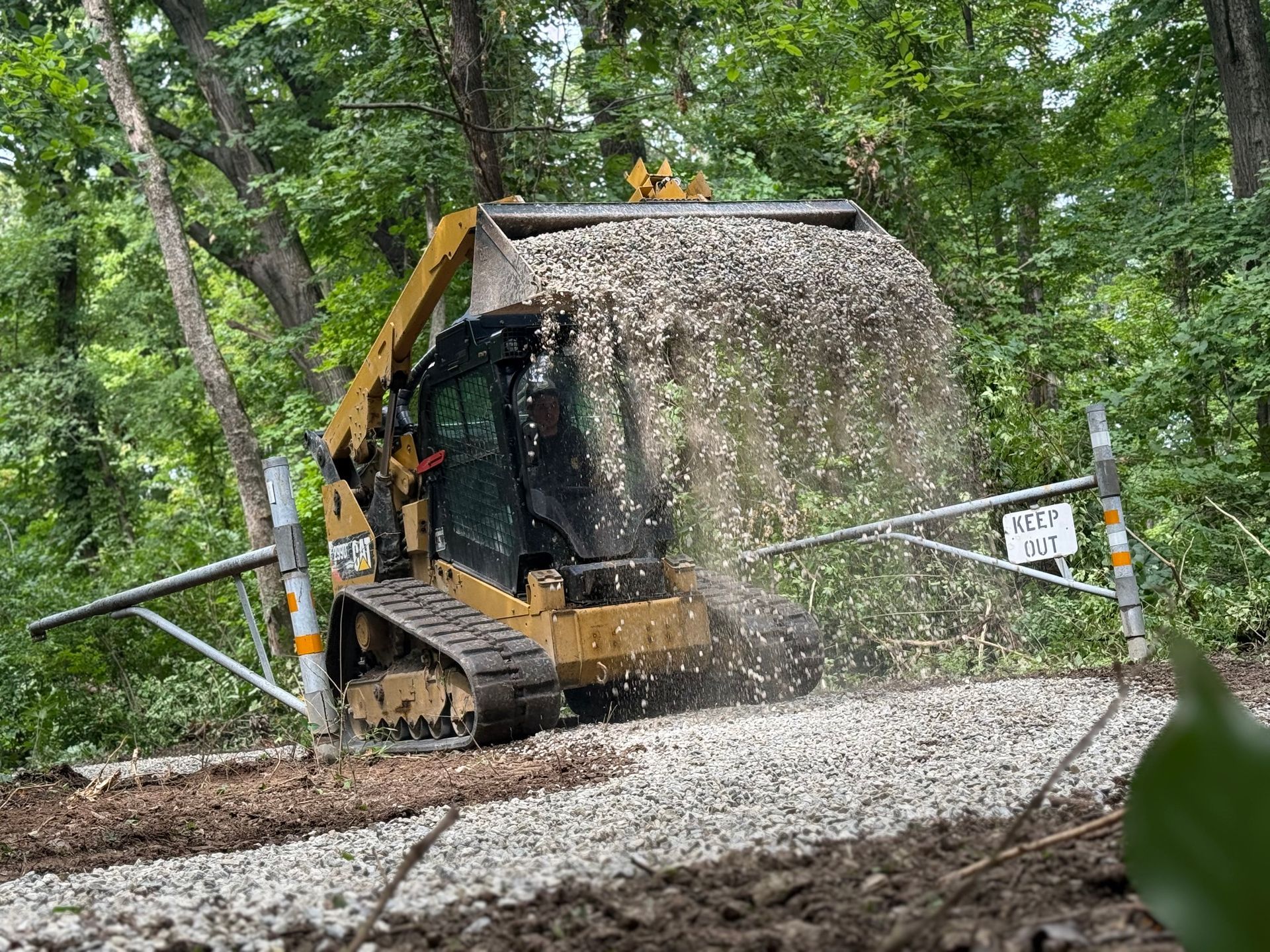 Yellow excavator covered in mud sprays gravel on a forest trail, with a metal gate in front.