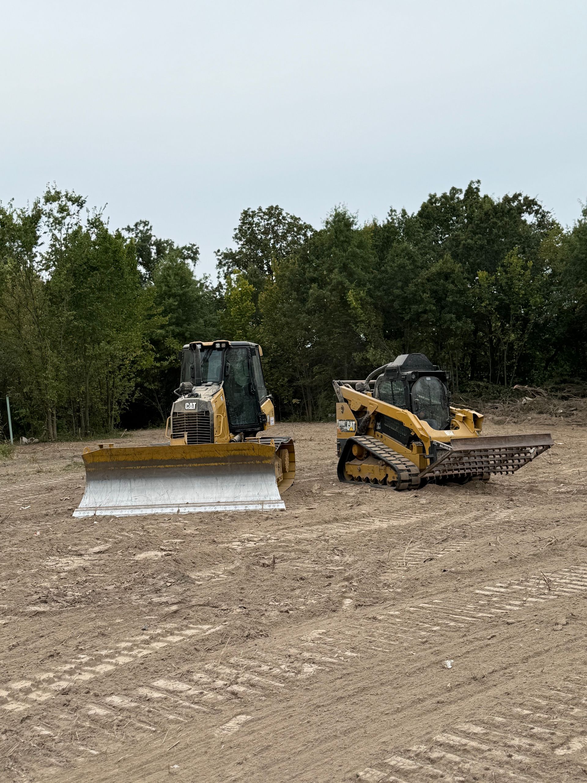 Two yellow bulldozers on a dirt lot, one pushing a blade, with trees in the background