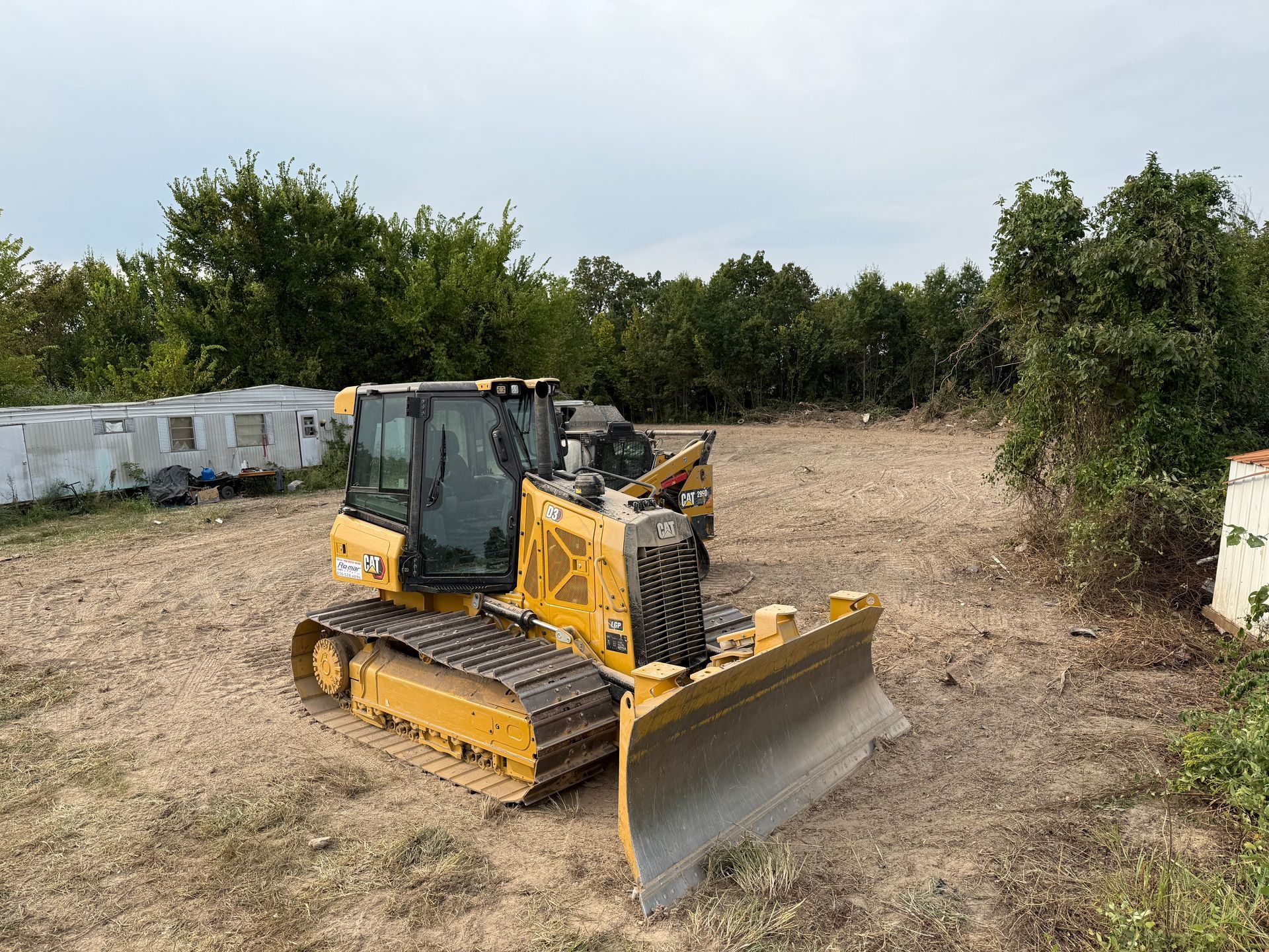 Yellow bulldozer leveling dirt in a cleared lot with trees and white barriers nearby