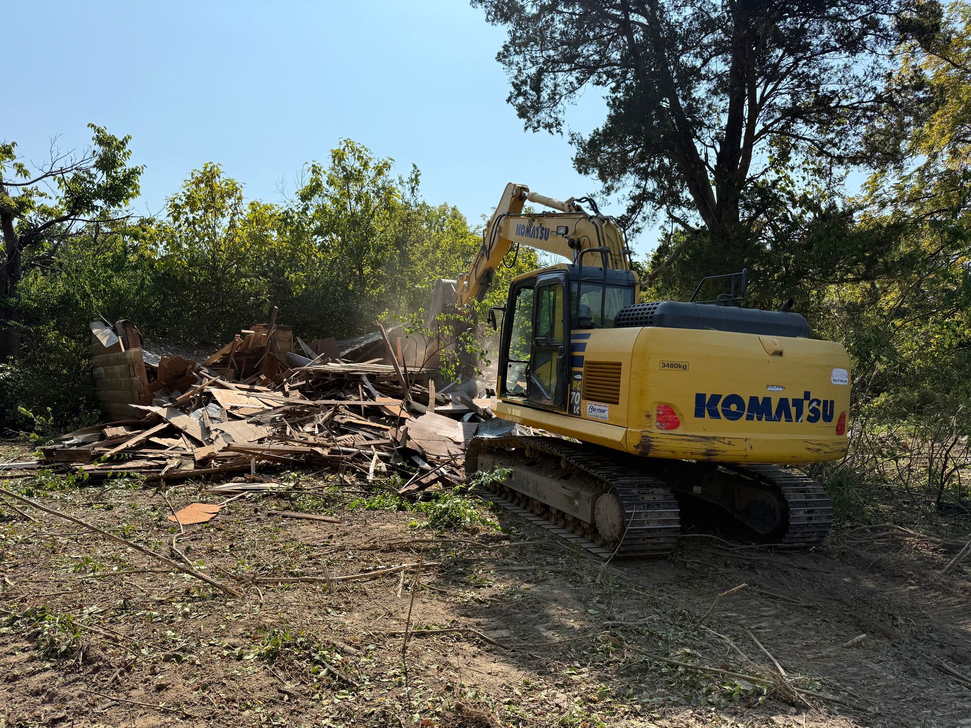 Yellow Komatsu excavator clearing a pile of debris beside trees on a sunny dirt lot