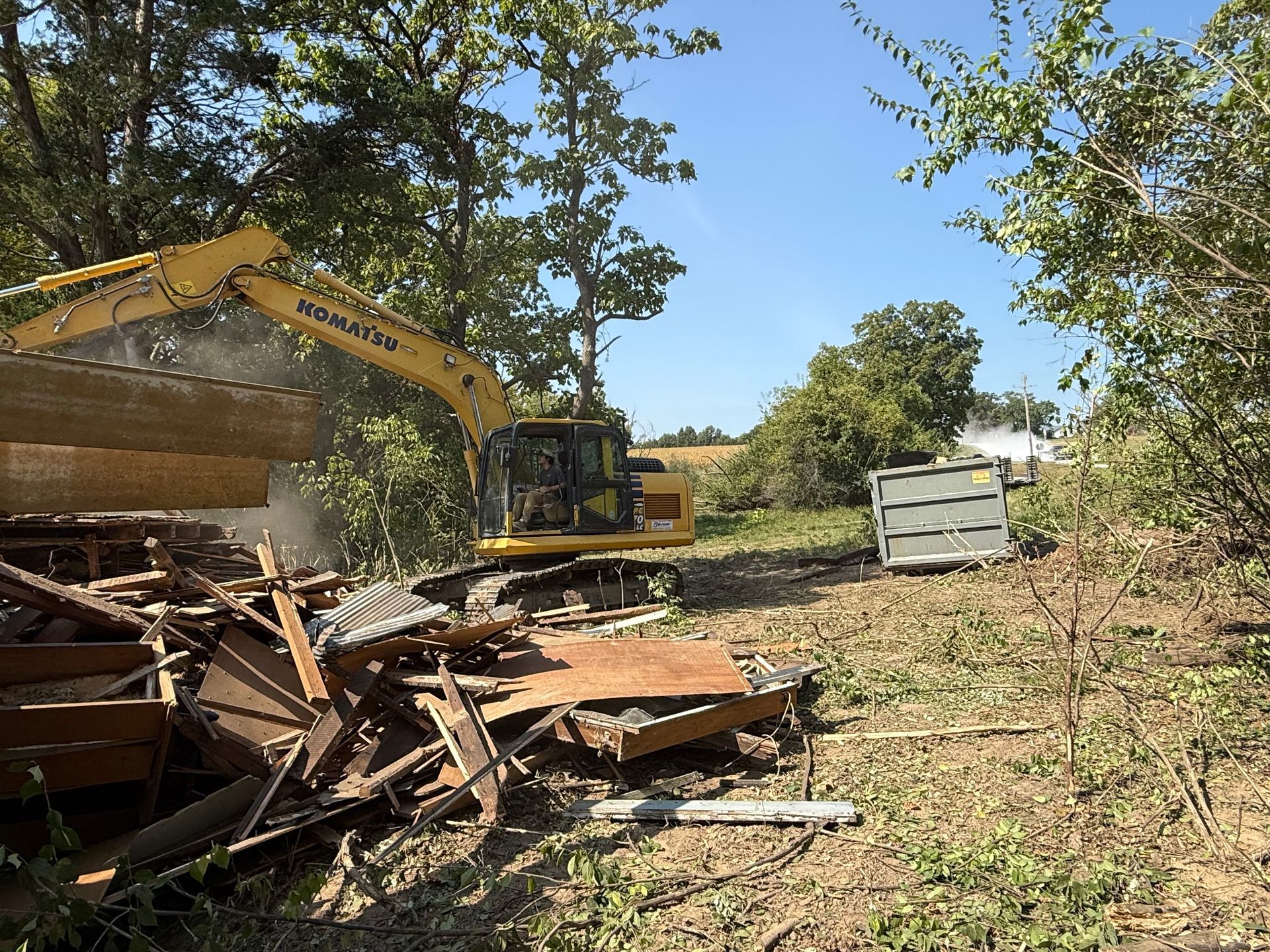 Excavator clearing debris beside a dirt path, with a white dumpster and trees in the background.