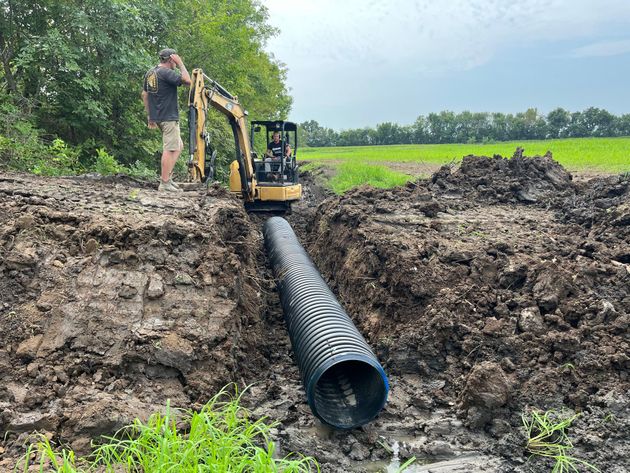 Excavator laying a large black drainage pipe in a muddy trench in a rural field