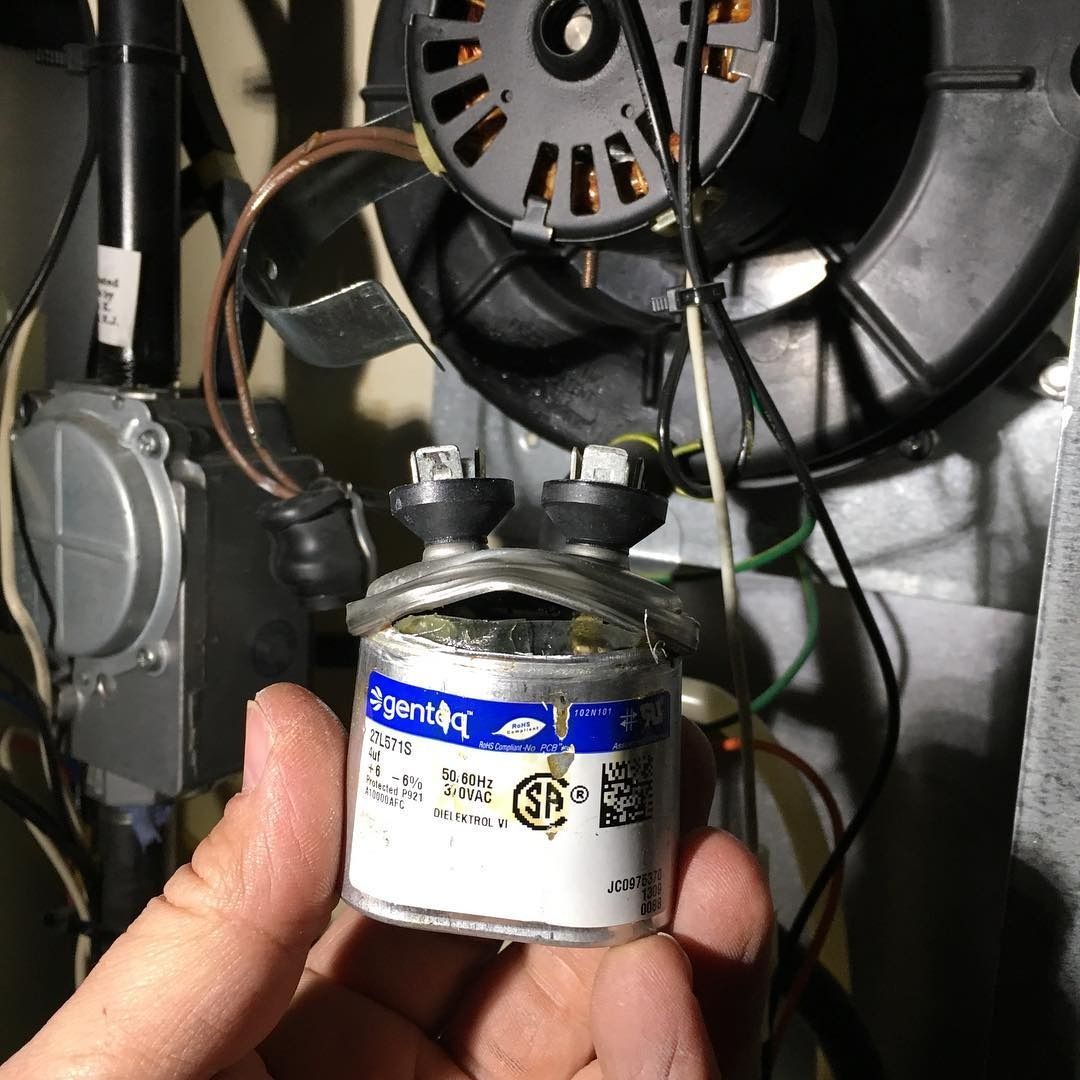 Hand holding a cylindrical capacitor next to a furnace blower fan.
