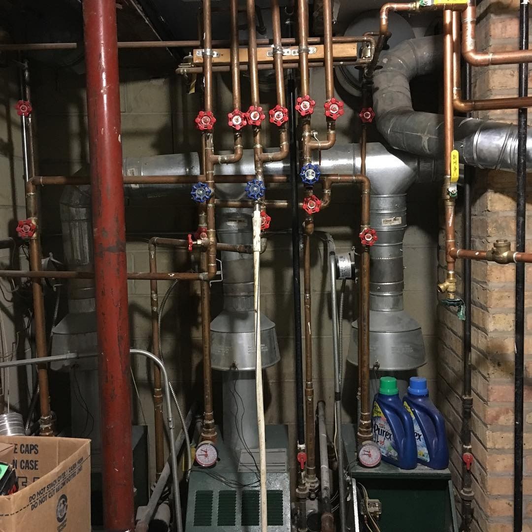 Copper pipes and valves against a brick wall, likely part of a heating system, with cleaning products on the floor.