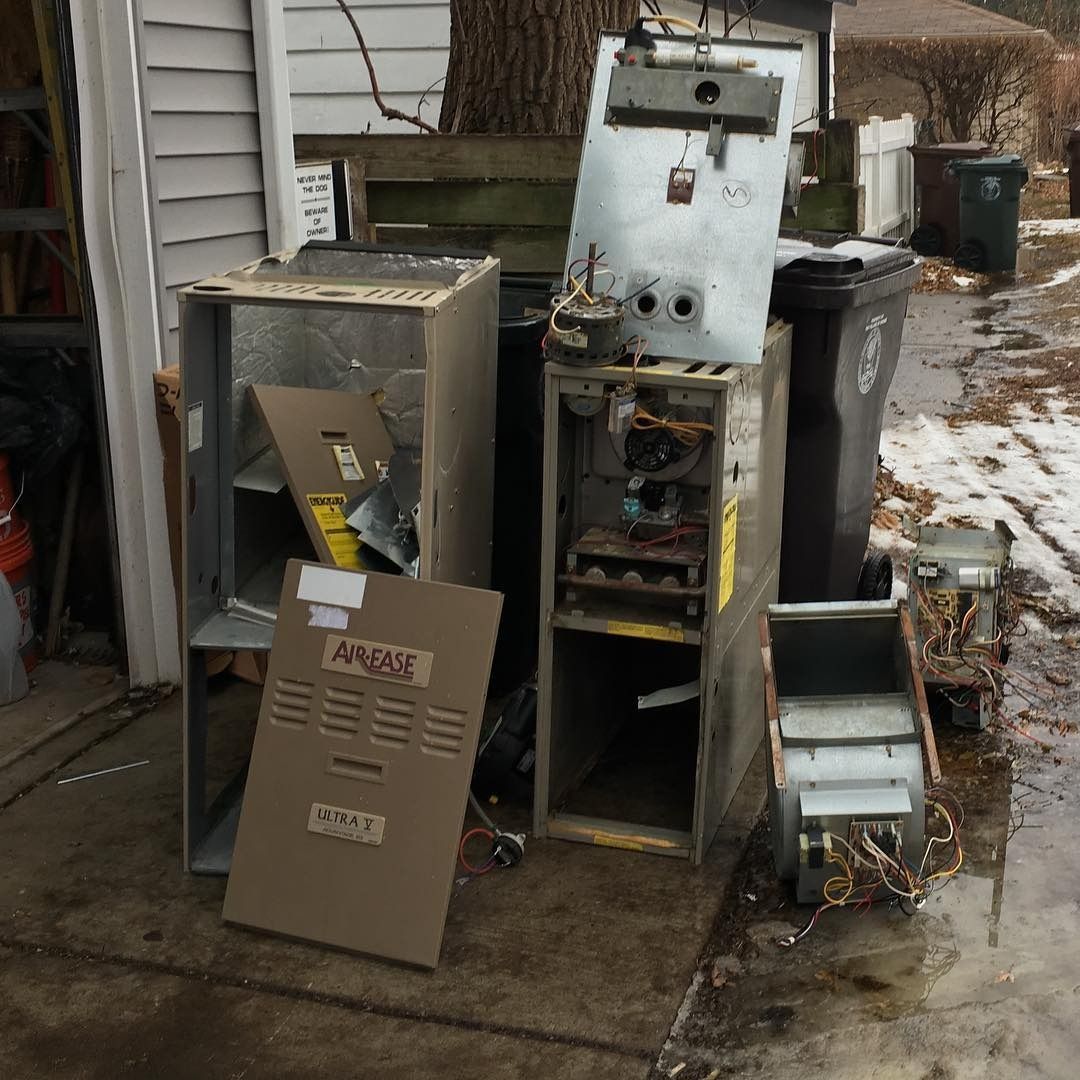 Broken furnace and water heater parts piled near a trash bin outdoors, likely for disposal.