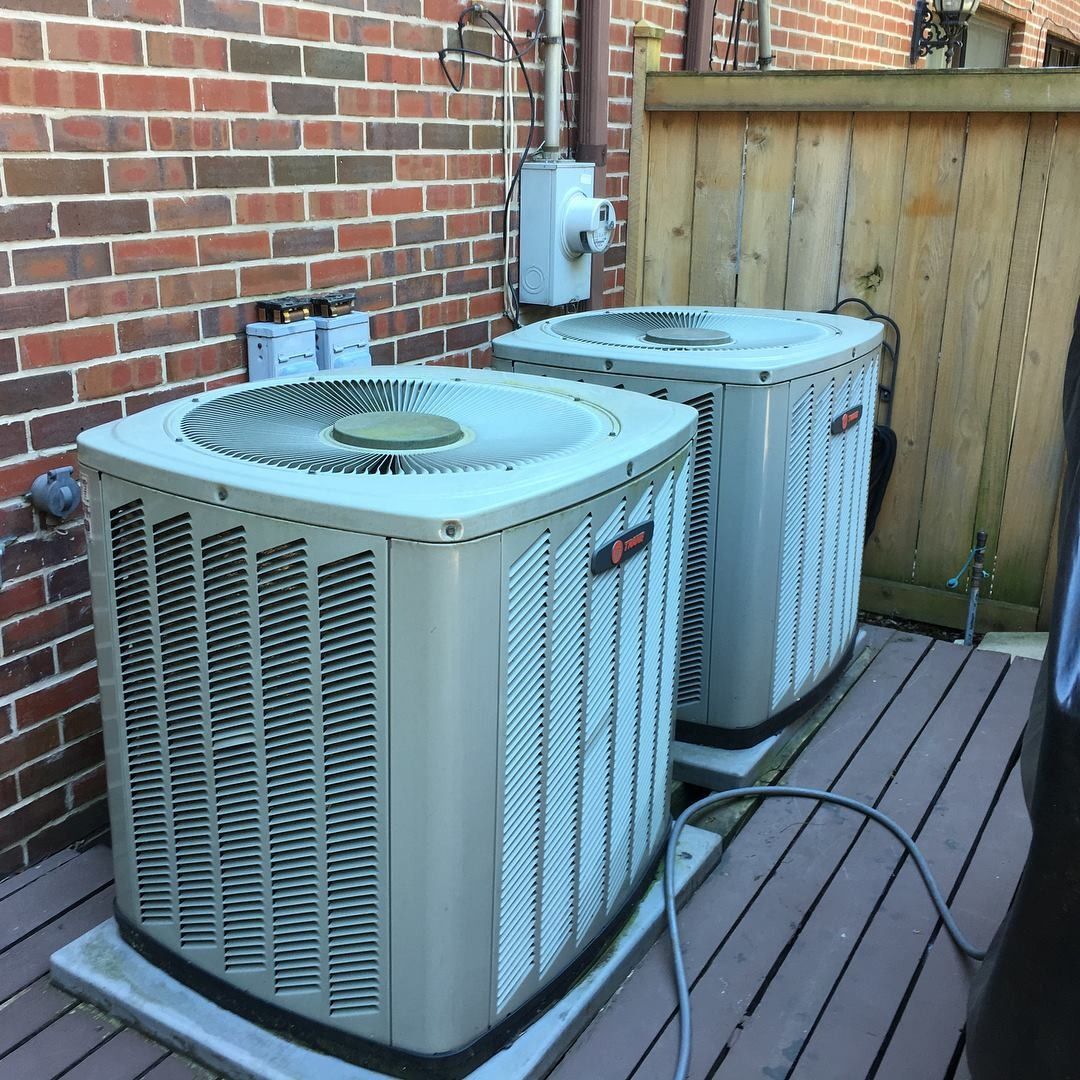 Two air conditioning units on a wooden deck next to a brick wall and wooden fence.