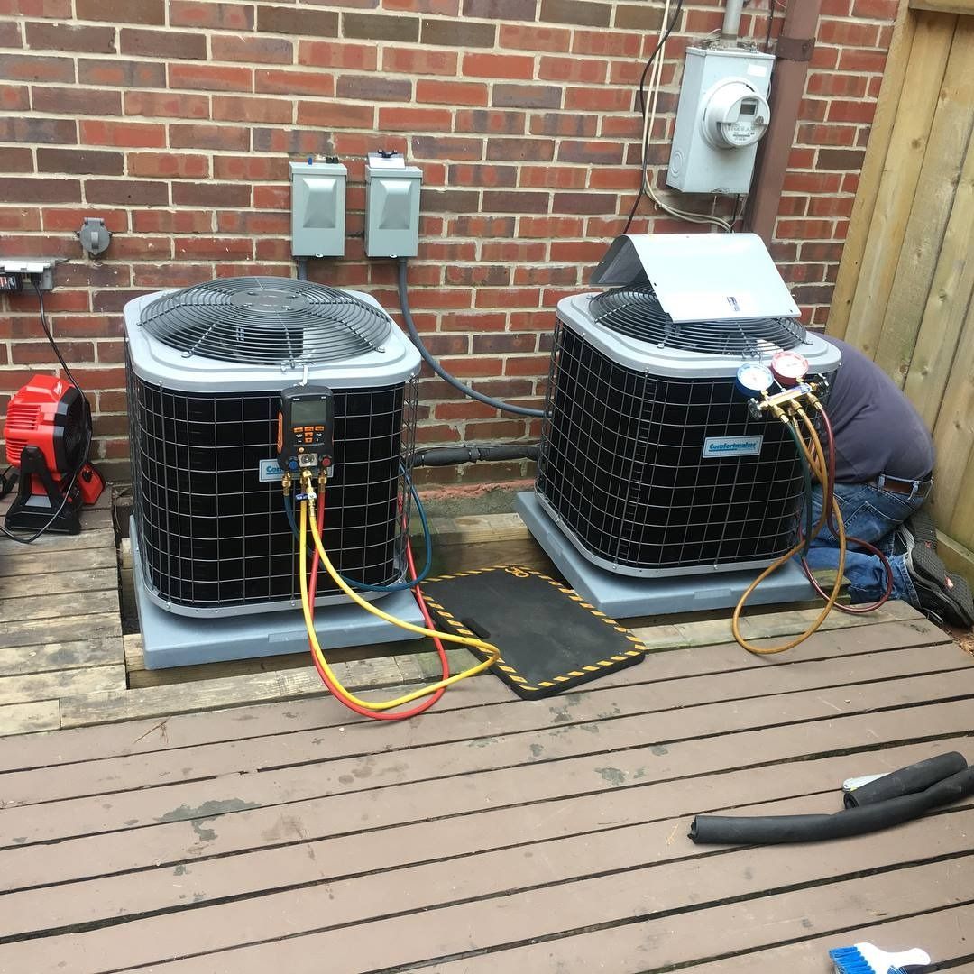 Two HVAC units on a wooden deck, with a person servicing one. Red brick wall in background.