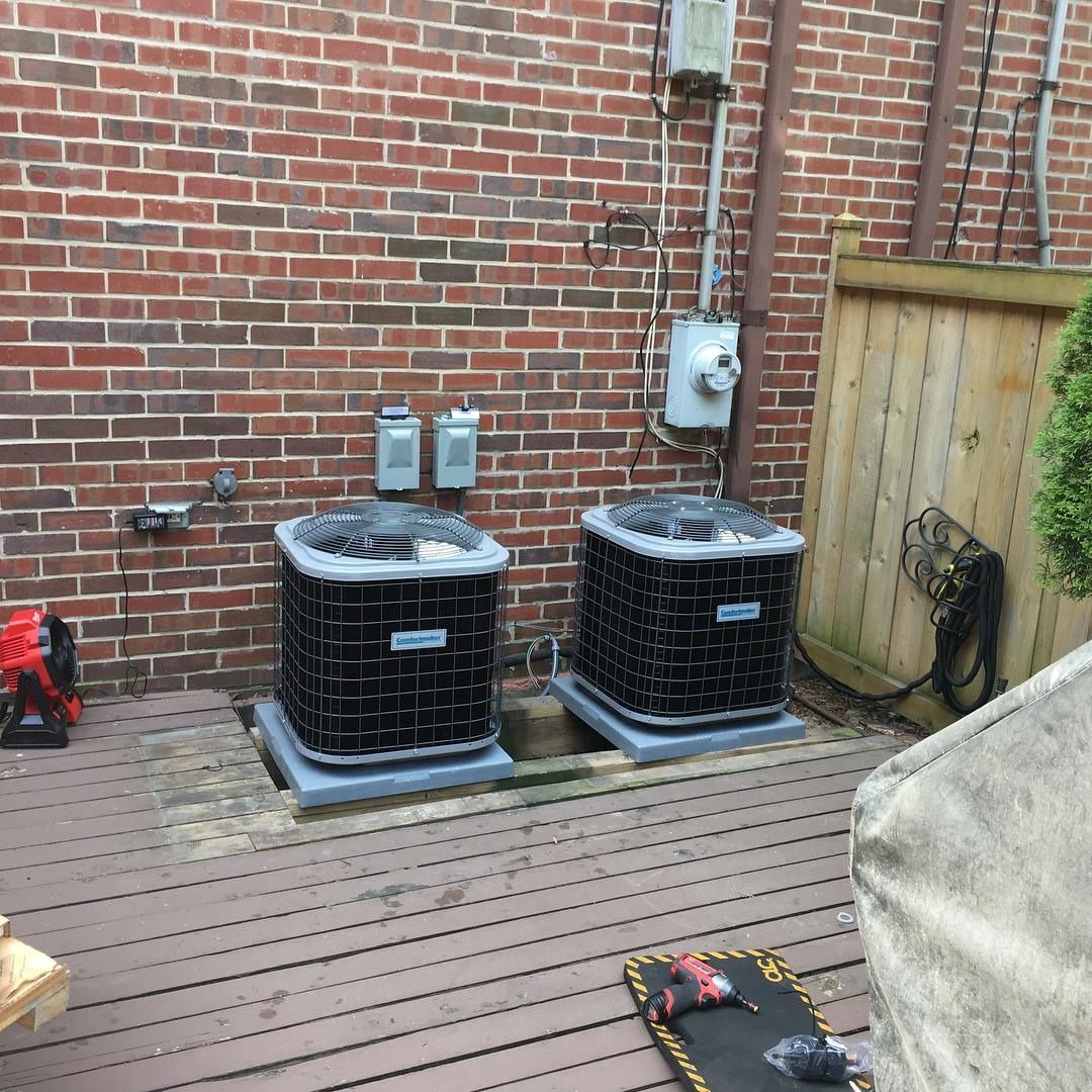 Two air conditioning units on a wooden deck next to a brick wall and a fence.