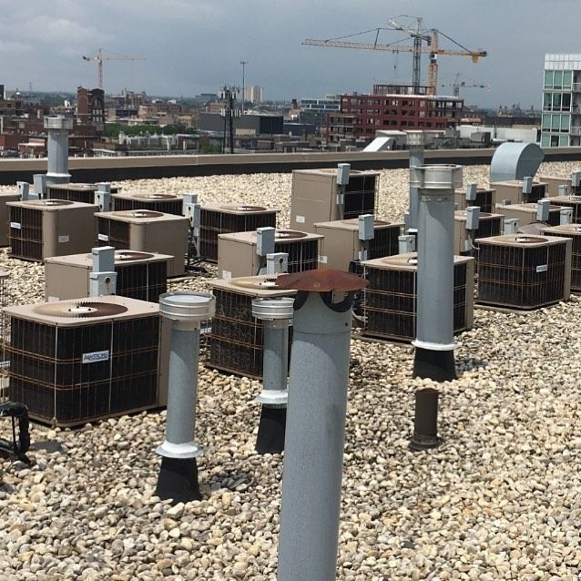 Rooftop with numerous air conditioning units and vertical pipes. Construction cranes and cityscape in the background.