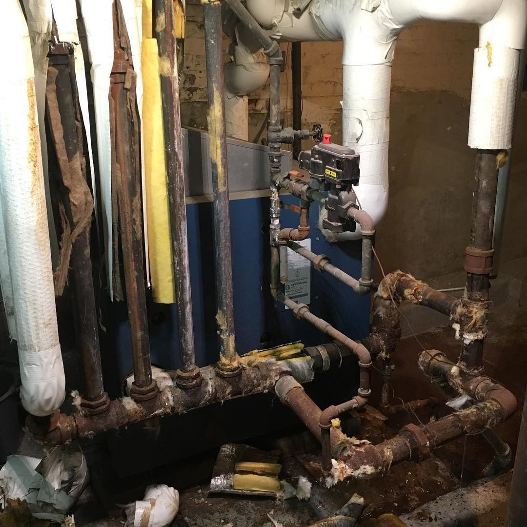 Old, rusty boiler with pipes, possibly in a basement. Some pipes have decaying insulation.
