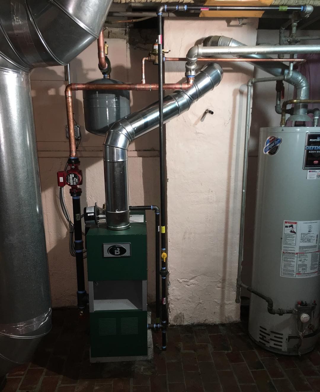 Green furnace and silver vent pipe in a basement setting with a water heater and pipes.