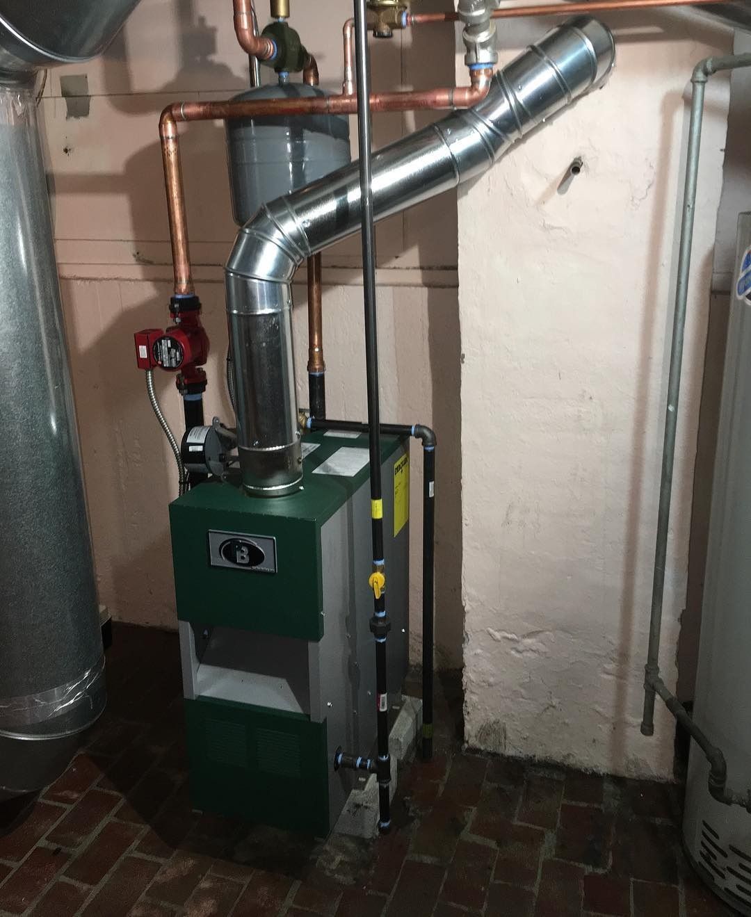 Green and silver furnace with copper pipes and a metal vent pipe in a basement.
