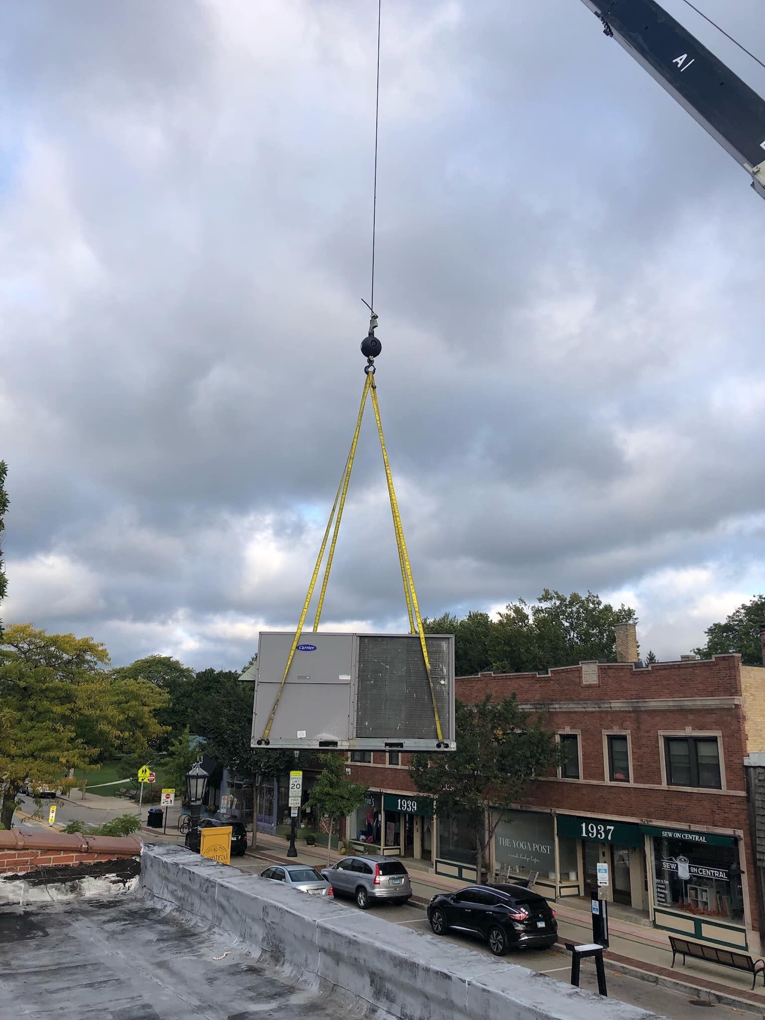 A crane lifts a building segment over a street with cars and buildings on a cloudy day.