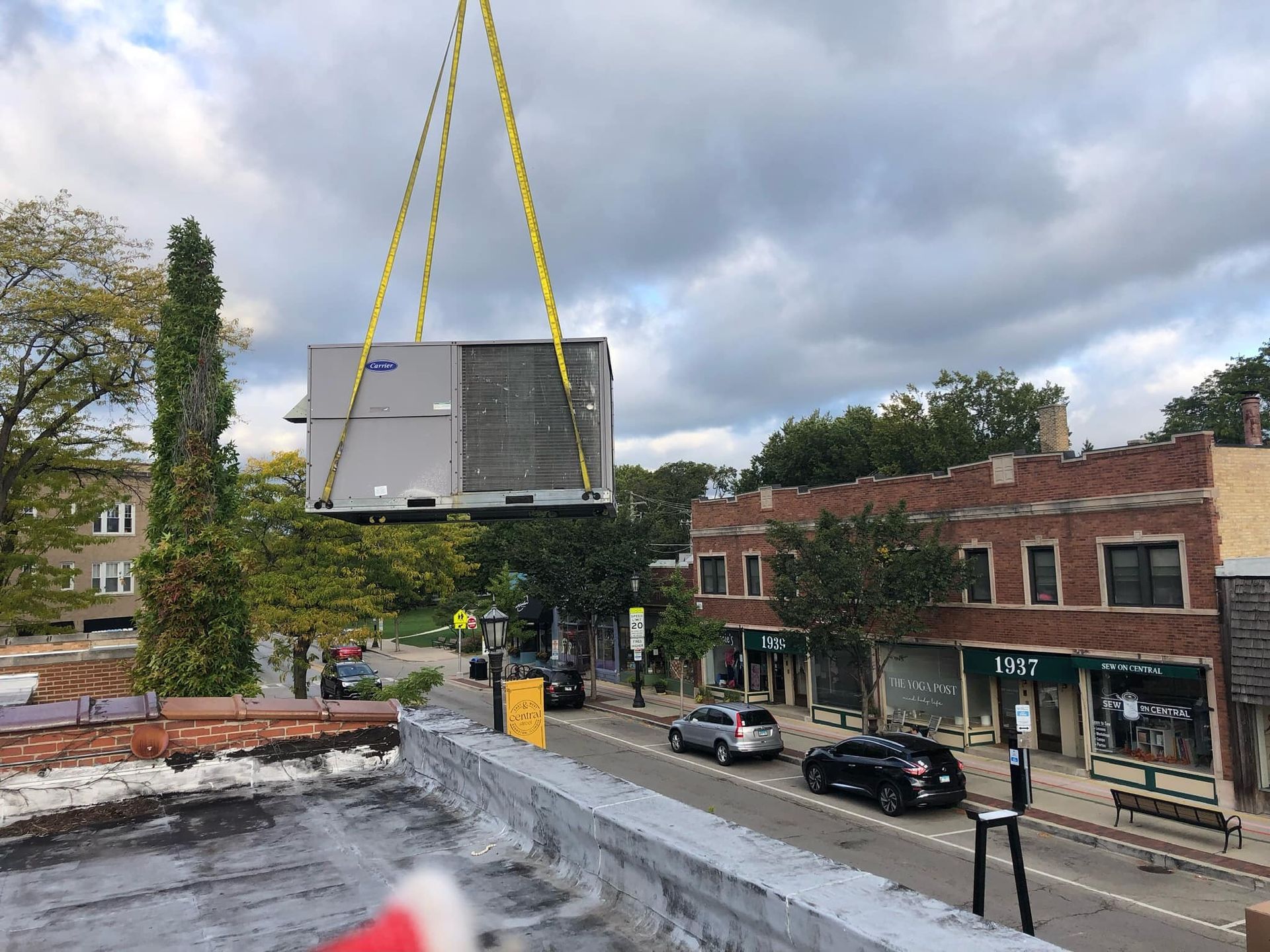 HVAC unit suspended by yellow chains over a street, being installed on a rooftop in a city setting.