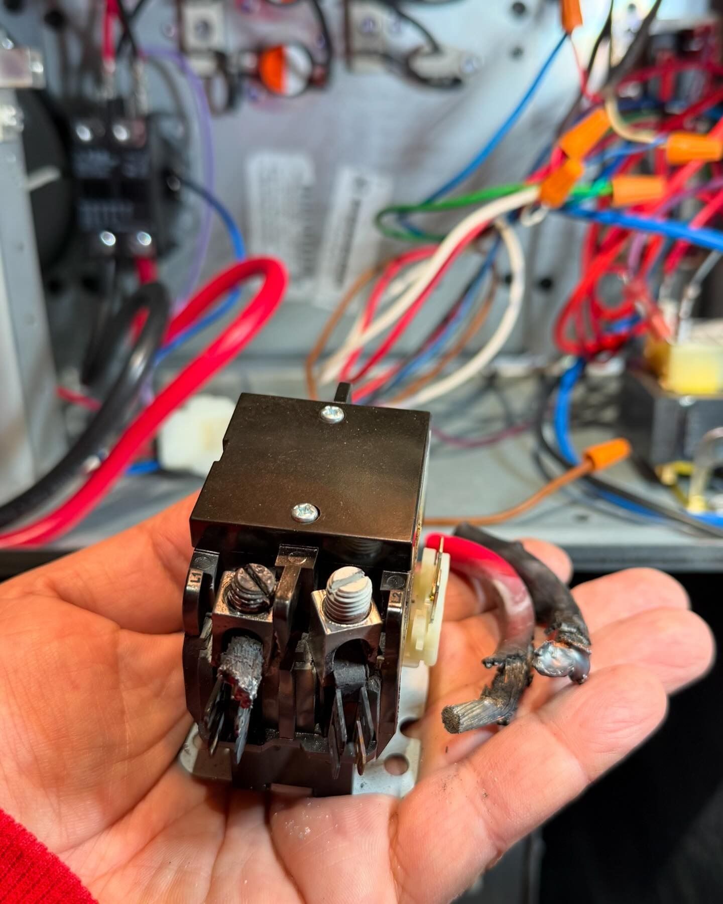 Hand holding a damaged black electrical contactor with burnt wires, in front of an open electrical panel.