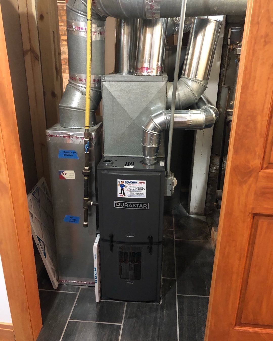 A furnace unit in a utility room. Gray and silver pipes connect to the unit. Wooden door frames border the space.