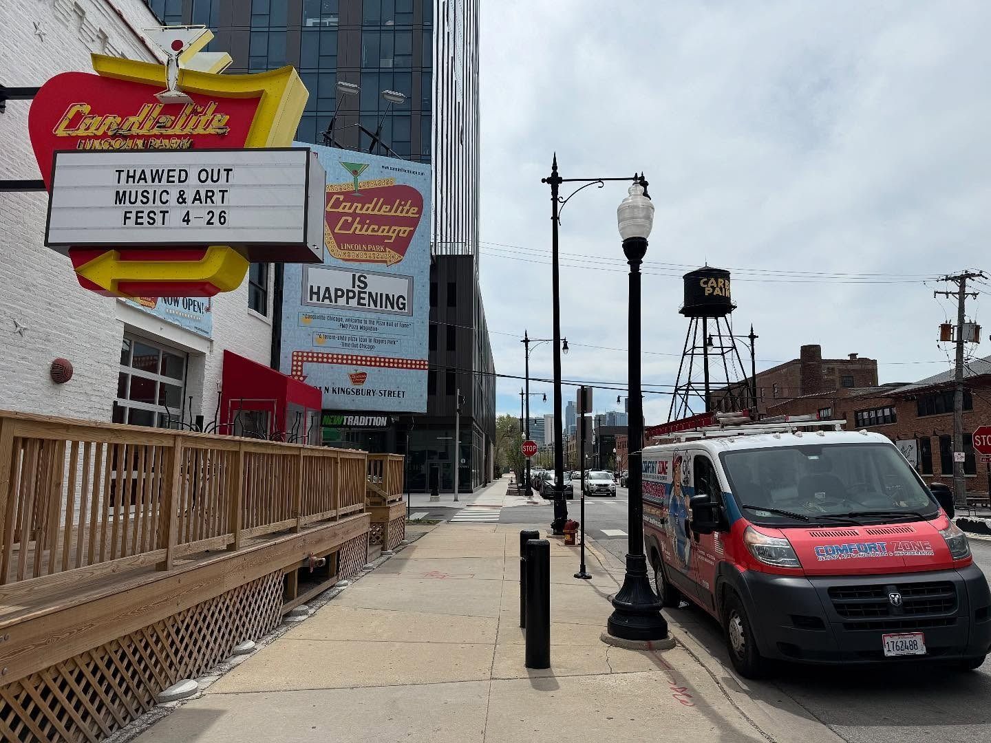 Street view with restaurant sign, building, and parked van. Wooden deck, sidewalk, and power lines.