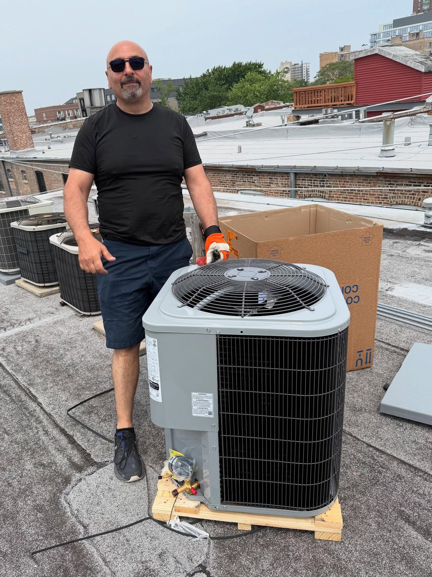 Man stands next to a new air conditioner on a rooftop, wearing gloves, shorts, and a black shirt.