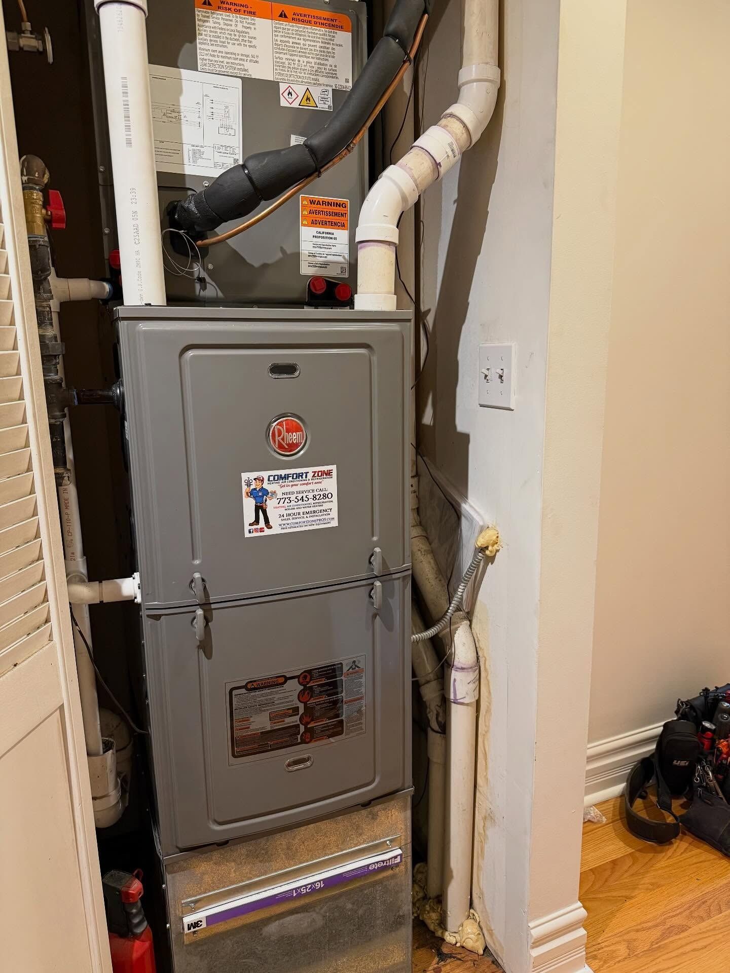 A Rheem furnace in a utility closet with white pipes and electrical outlet.