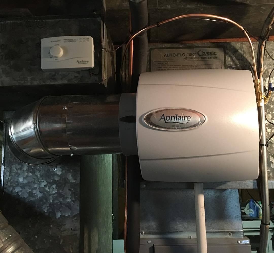 Aprilaire humidifier attached to ductwork in a basement. White unit with silver trim, copper tubing.