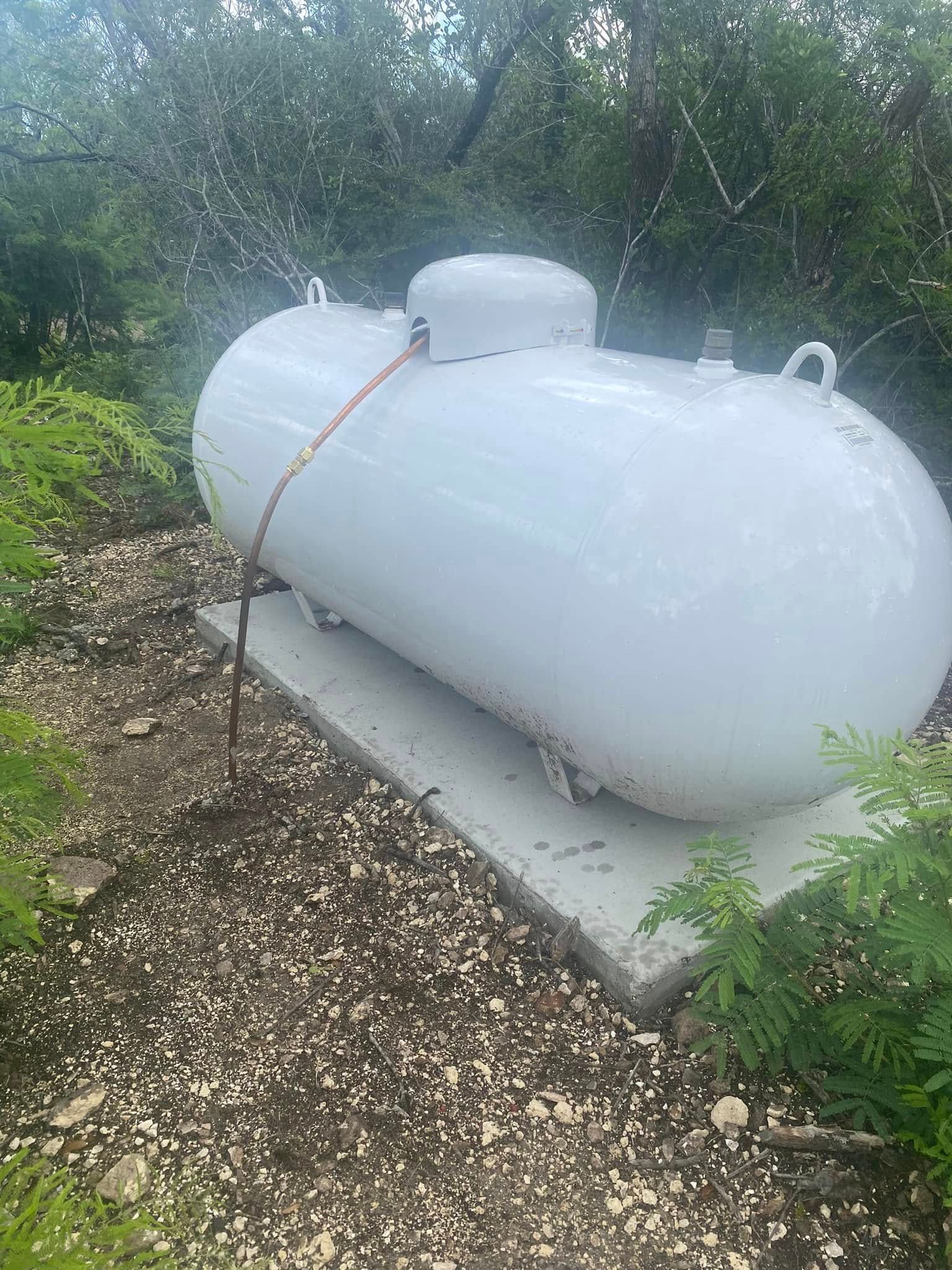 White cylindrical propane tank on a concrete pad, surrounded by greenery and wet ground.