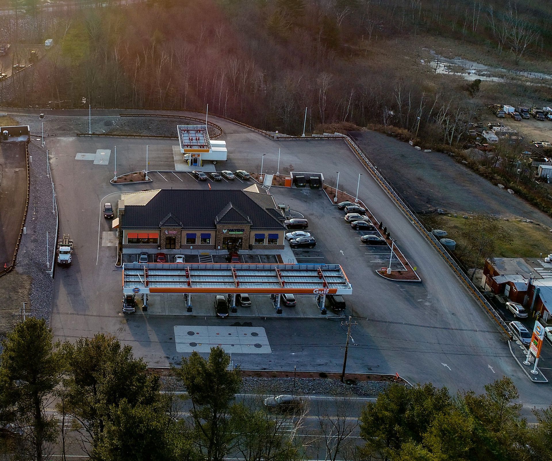 Aerial view of a gas station and convenience store with a large parking area, set against a wooded landscape.