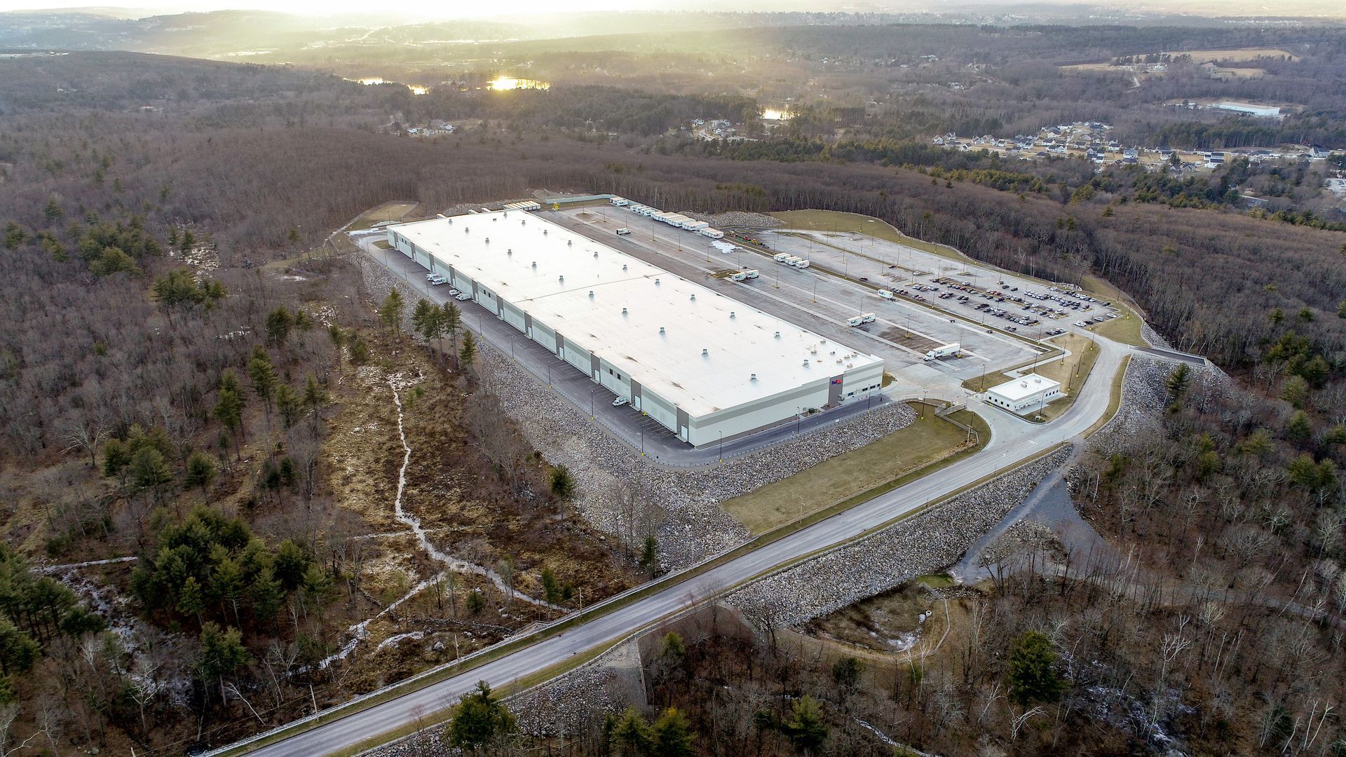 Aerial view of a long, white-roofed industrial warehouse building surrounded by a parking lot and dense forest.