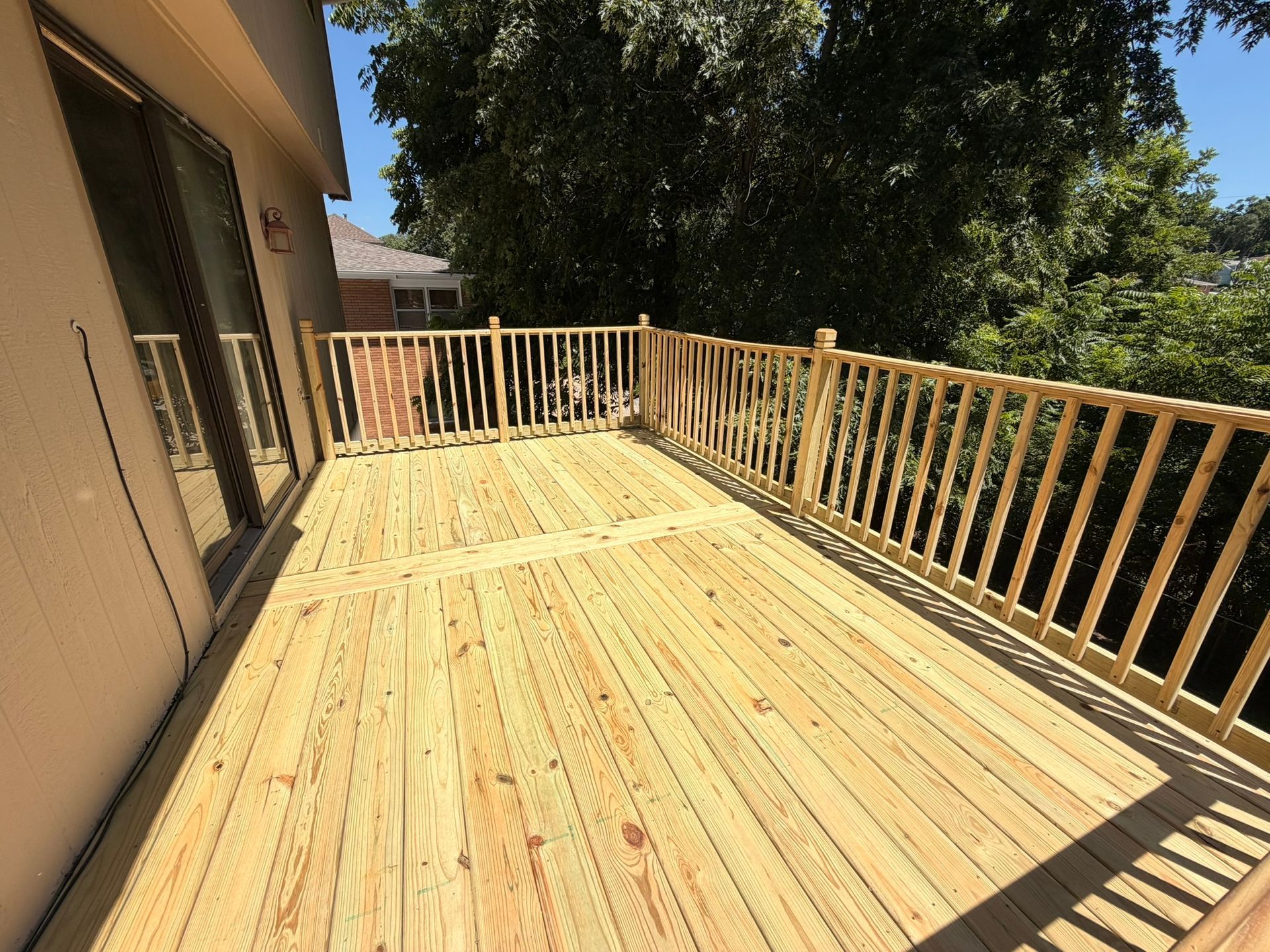 Sunlit wooden deck with railings beside a house, overlooking trees and yard