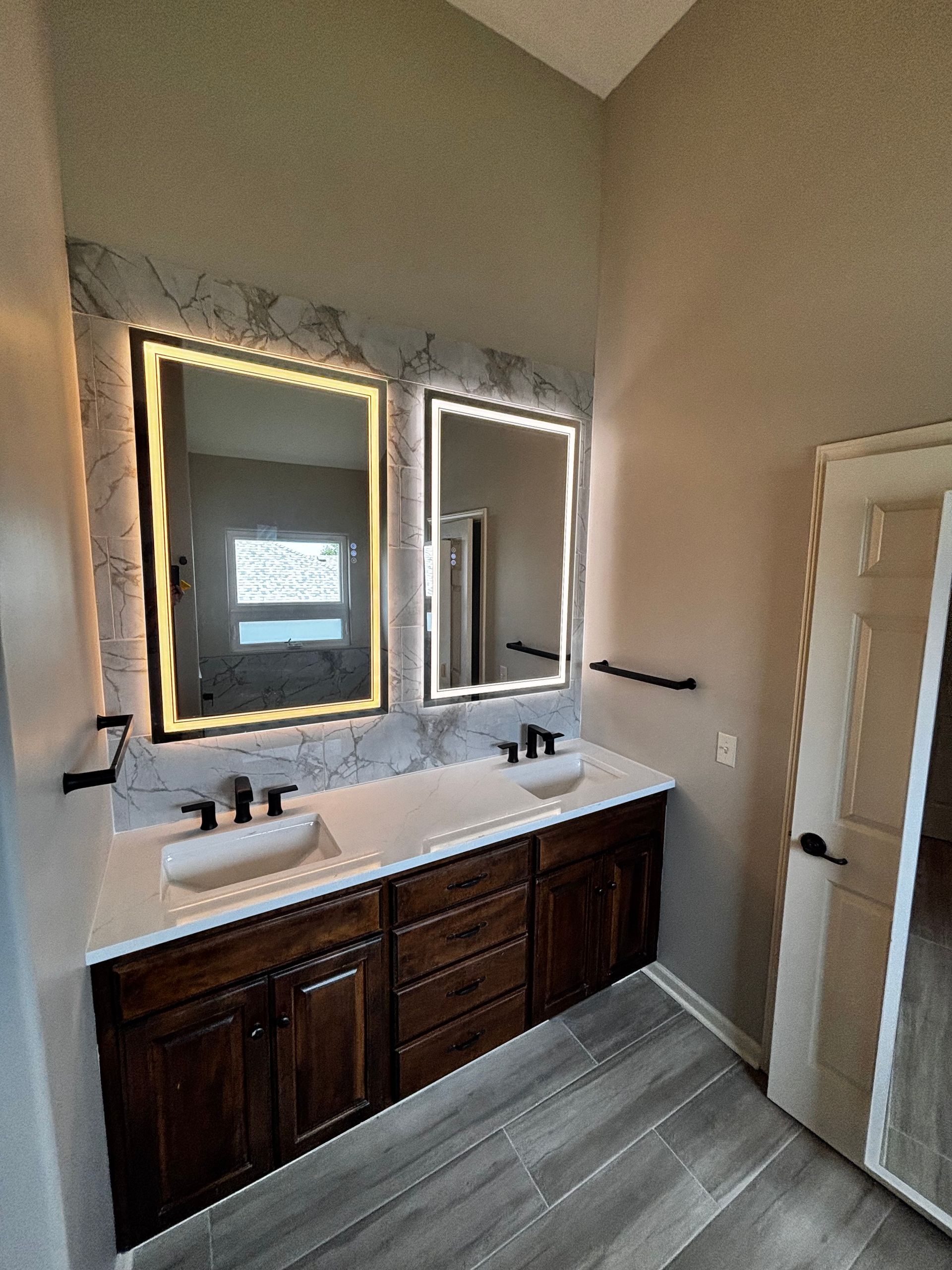 Bathroom with dual sinks, dark brown vanity, two illuminated mirrors, and light-colored flooring. White countertop, black hardware, and a tall cabinet on the right.