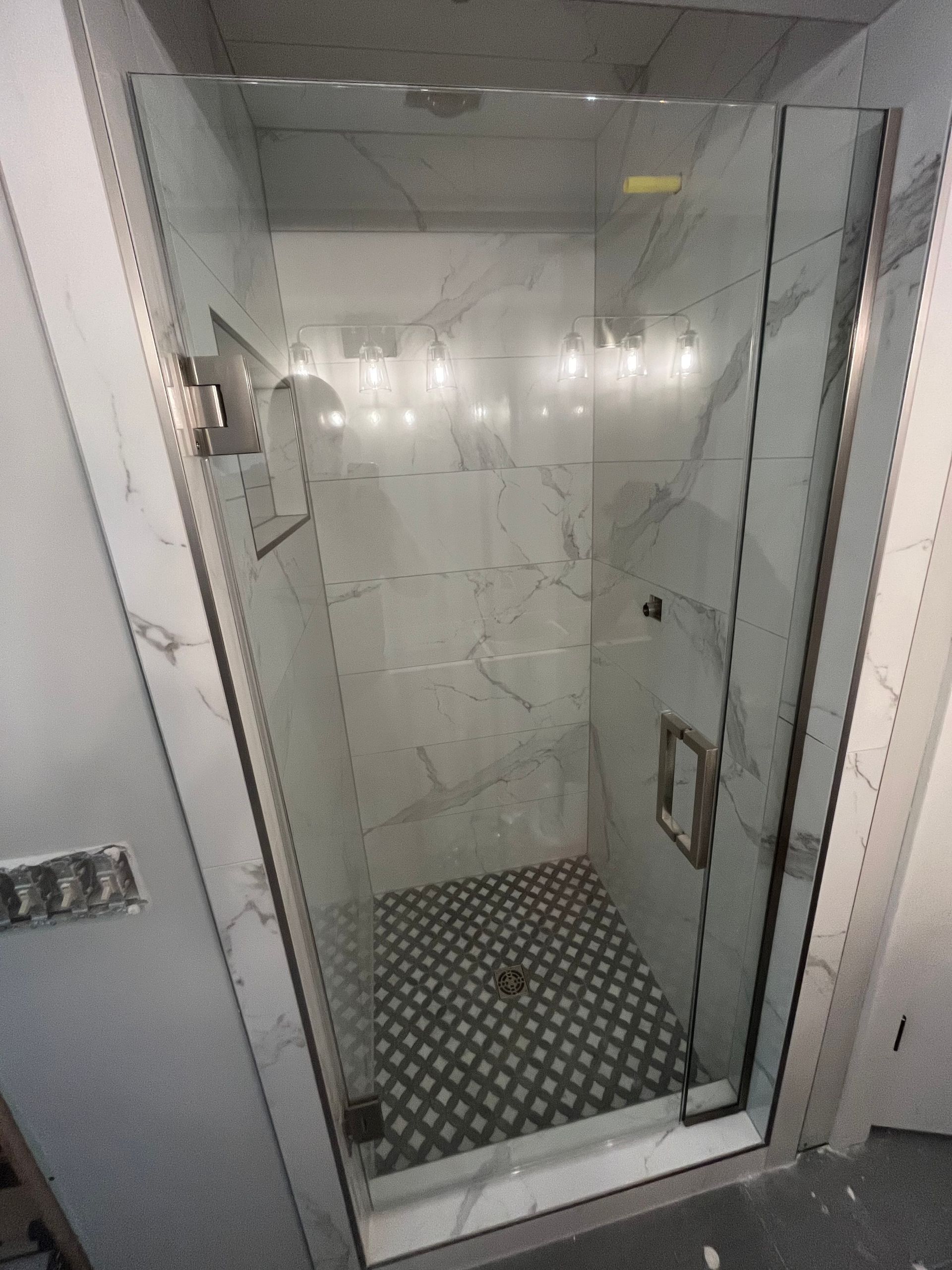 A modern shower with a glass door. White marble-like tiles line the walls, contrasting with the dark tiled floor.