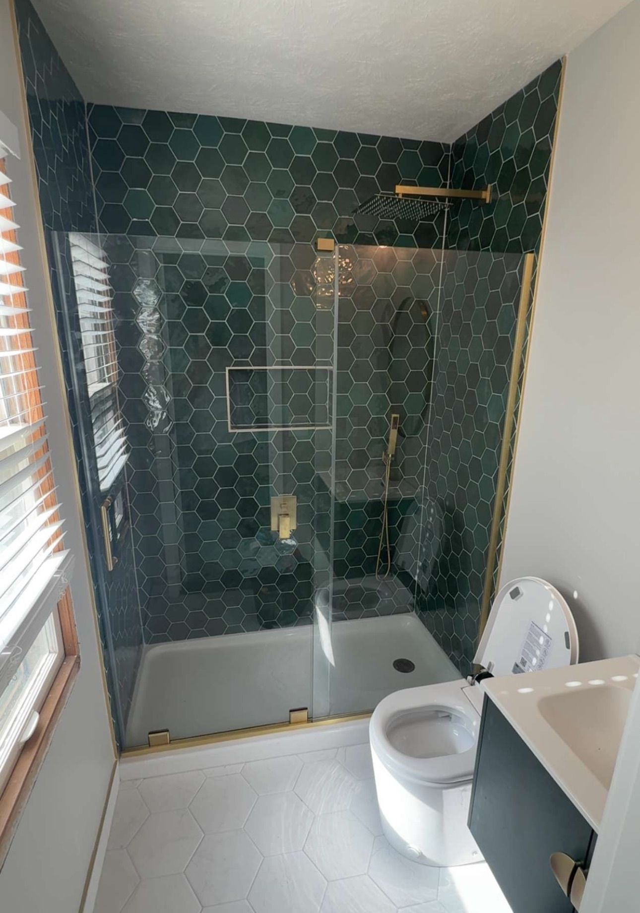 Bathroom with emerald green hexagon tile shower, gold fixtures, and a glass door. White toilet, vanity, and light-colored floor.