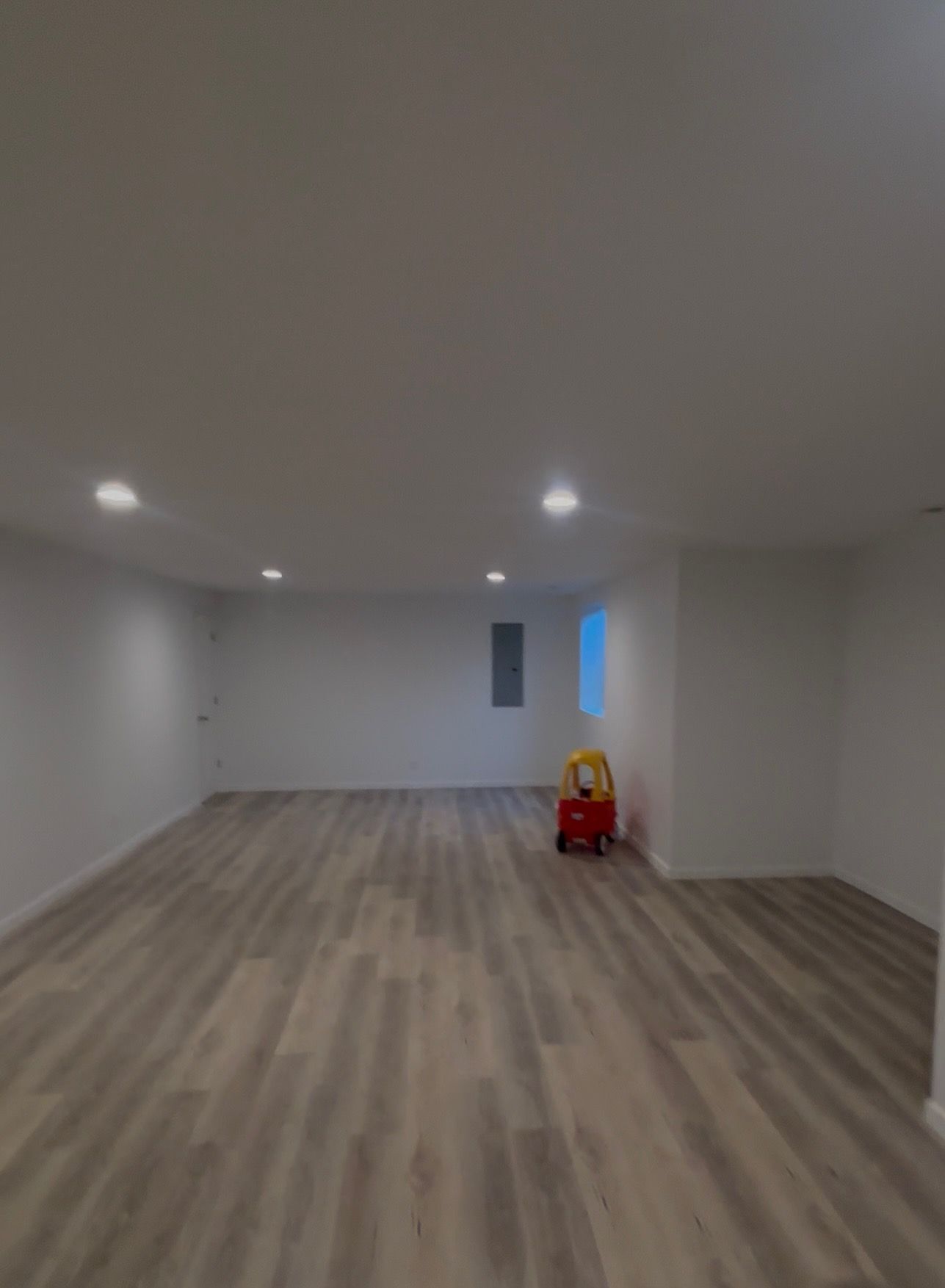 Empty basement room with wood-look flooring, white walls and ceiling. Small red wagon toy sits against a wall.