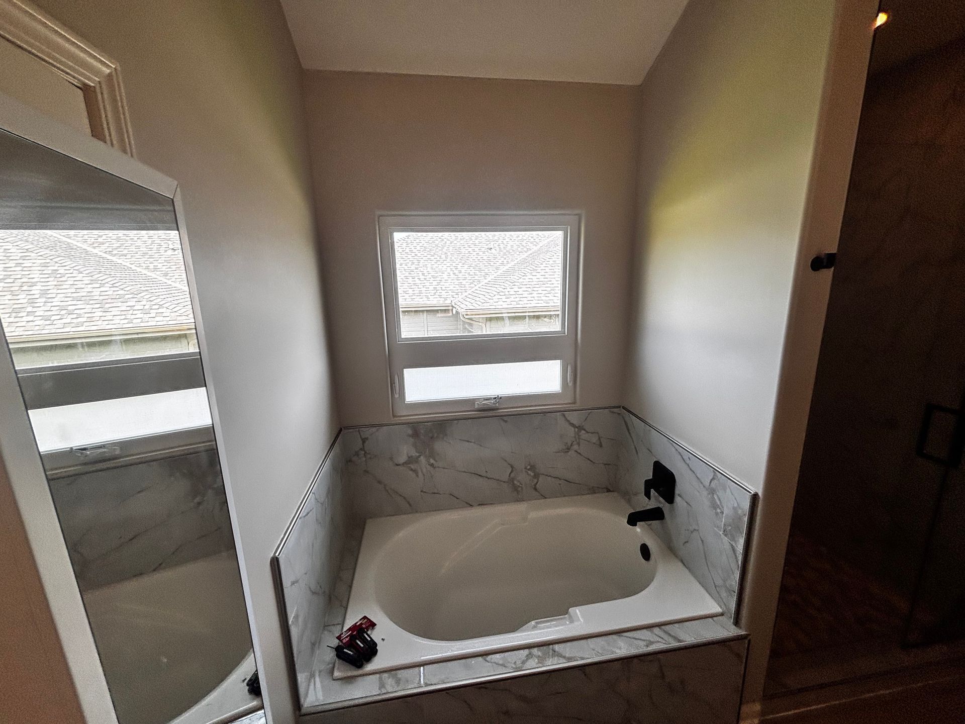 Bathroom with a white bathtub surrounded by marble tile, a window above it, and a mirror on the left.