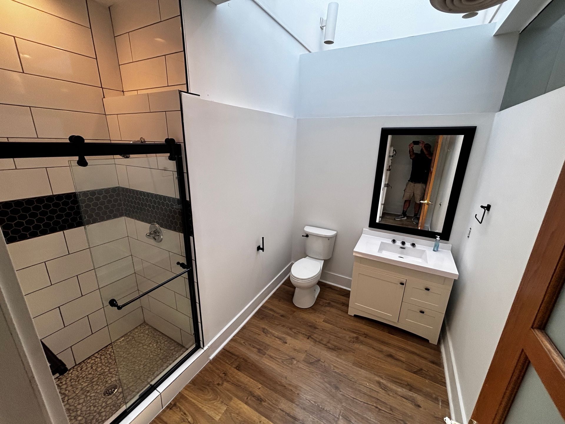 Bathroom with shower, toilet, and vanity. White walls, dark trim, wood-look flooring. Black framed shower door.