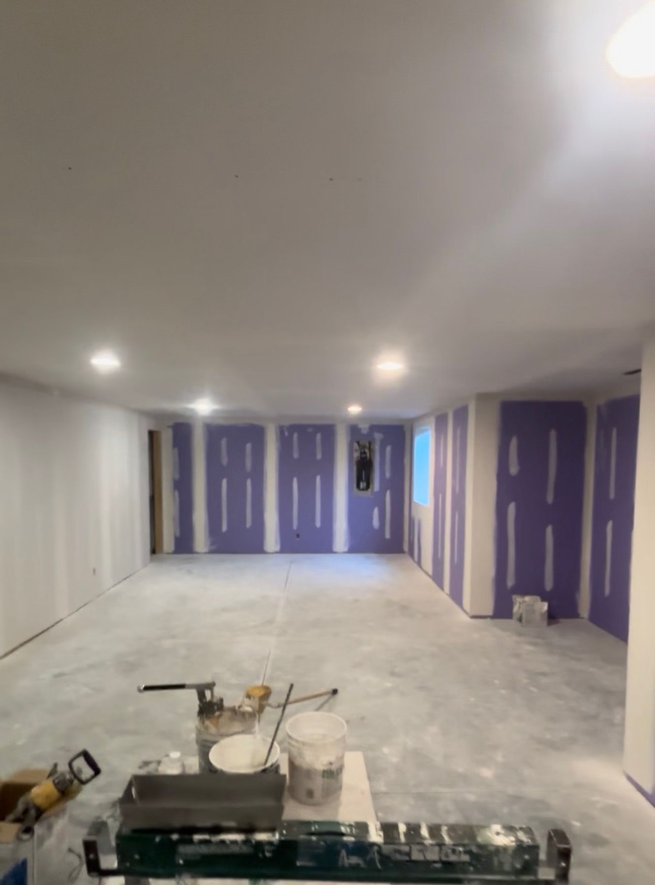 A basement room under construction. Purple drywall lines walls. Paint supplies rest on the concrete floor.