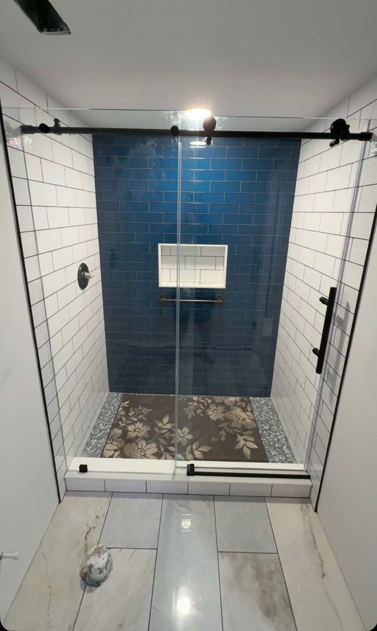 Modern shower with black-framed glass door, blue tiled accent wall, white tiled walls, and floral rug.