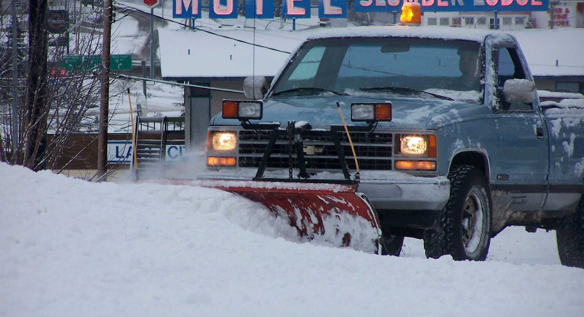 A blue pickup truck with an attached red snowplow clears a snow-covered road in front of a motel.