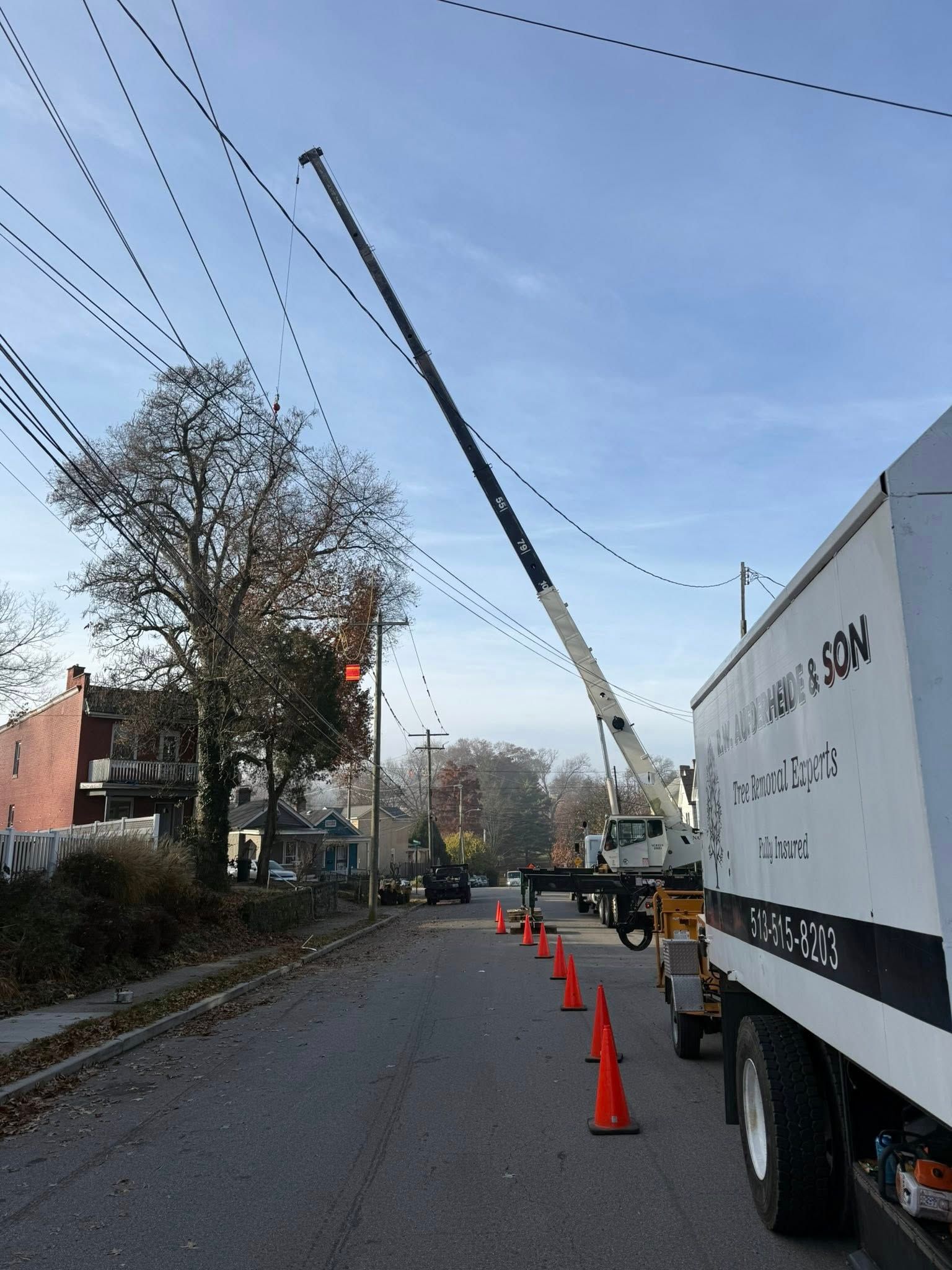 A utility truck with boom extending towards power lines on a street with orange cones.