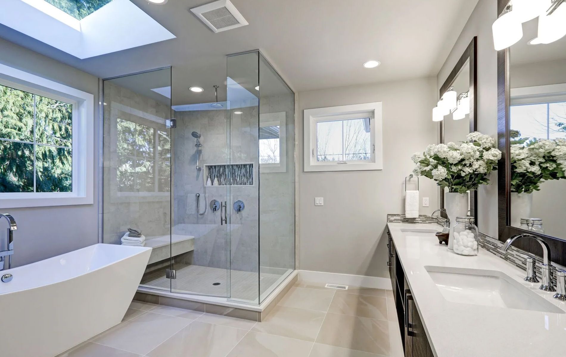 Modern bathroom with glass shower, soaking tub, double vanity, and skylight. Neutral colors.