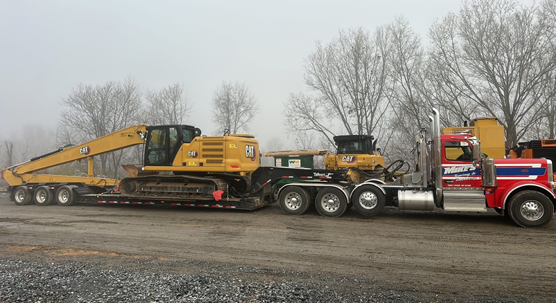 Yellow excavator and smaller machine on a flatbed trailer pulled by a red semi-truck. Trees in the background.