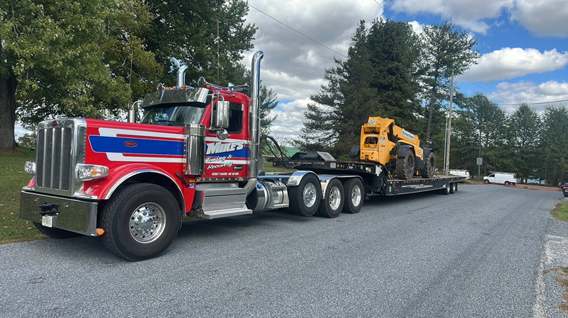 Red semi-truck with a flatbed trailer transporting a yellow skid steer on a gravel road, trees in the background.