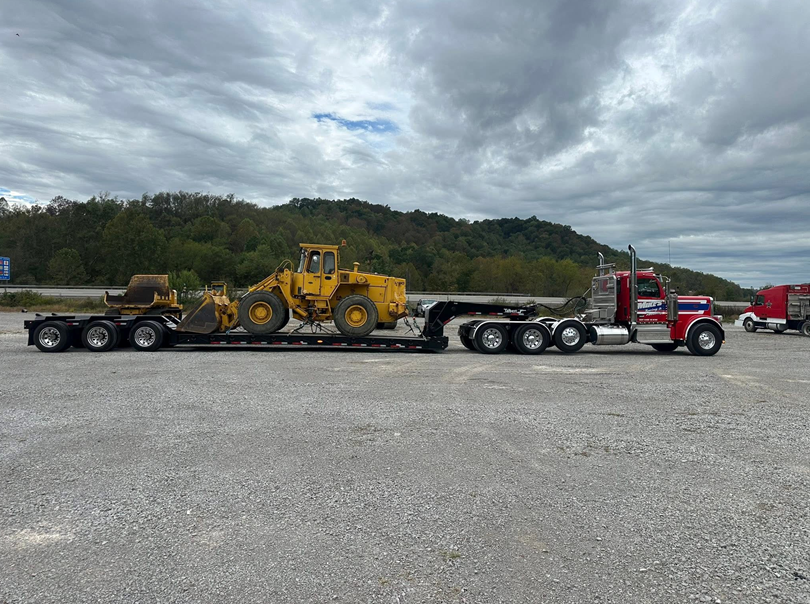 Semi-truck hauling yellow construction equipment on a flatbed trailer, parked on asphalt under cloudy skies.