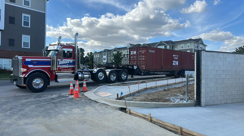 Semi-truck hauling a shipping container in front of an apartment building.