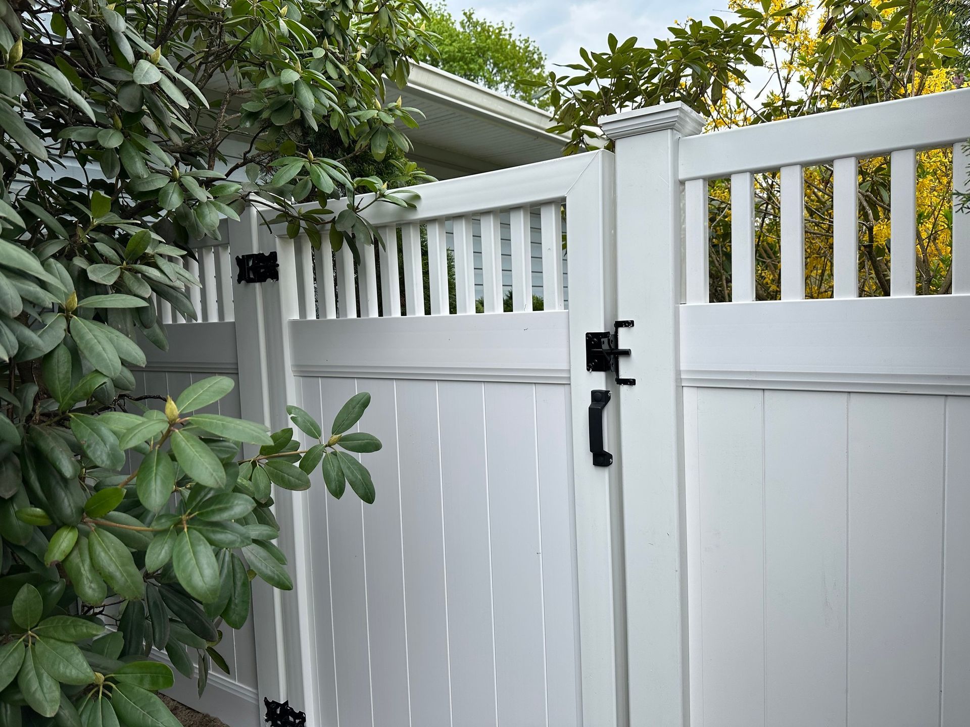 White gate with black hardware and vertical slats, framed by green foliage.