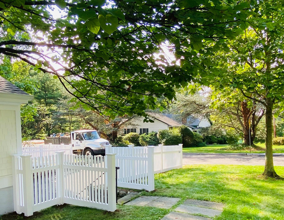 White picket fence in front of a house with a white vehicle parked behind it, under a sunny tree canopy.