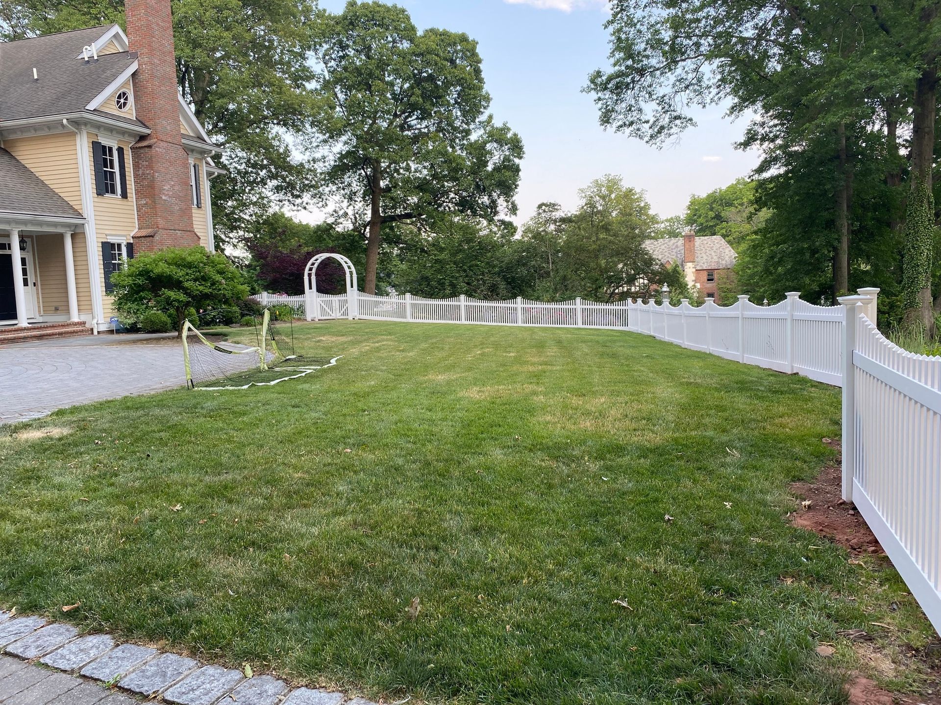 Lawn with white picket fence, stone driveway, and yellow house in the background under a blue sky.